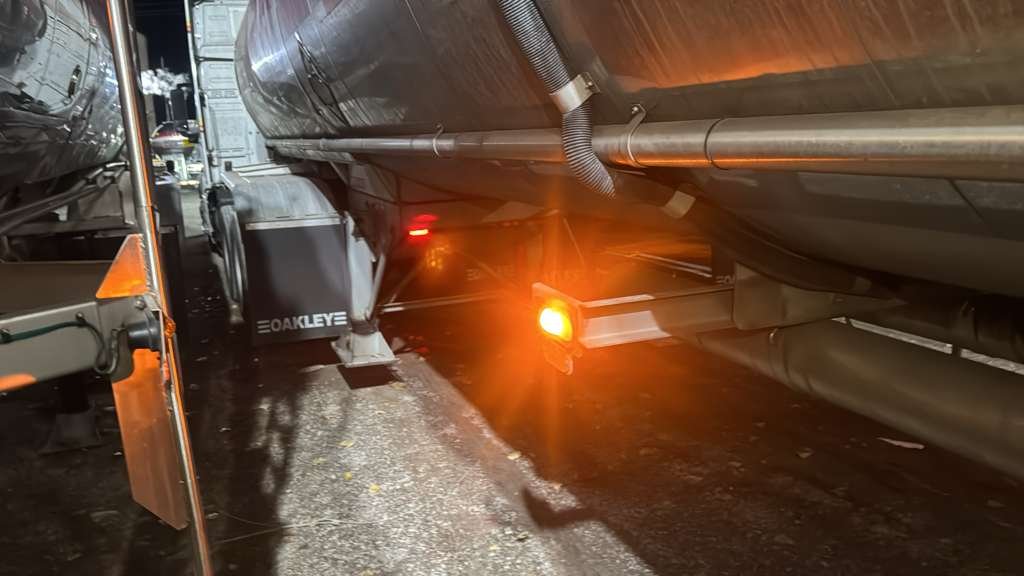 Close-up view of the underside of a large tanker truck, showing pipes, a fuel tank labeled 'OAKLEY', and safety reflectors with lights.