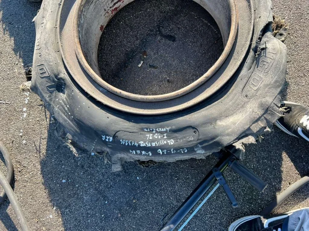 Large used tire lying on the ground, with visible white markings and writing on it, surrounded by asphalt, part of a bicycle and other shoes nearby.