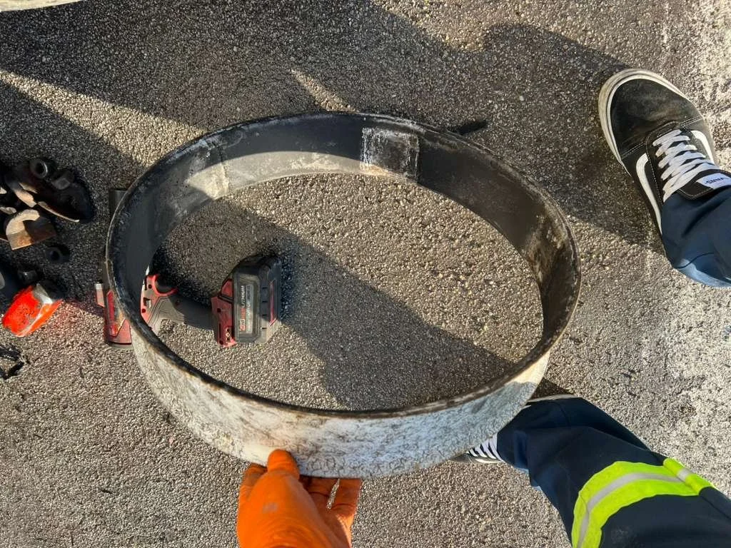 Person holding a dirty, circular metal band or ring on the ground, with tools around and a person wearing a safety vest standing nearby on a textured asphalt surface.