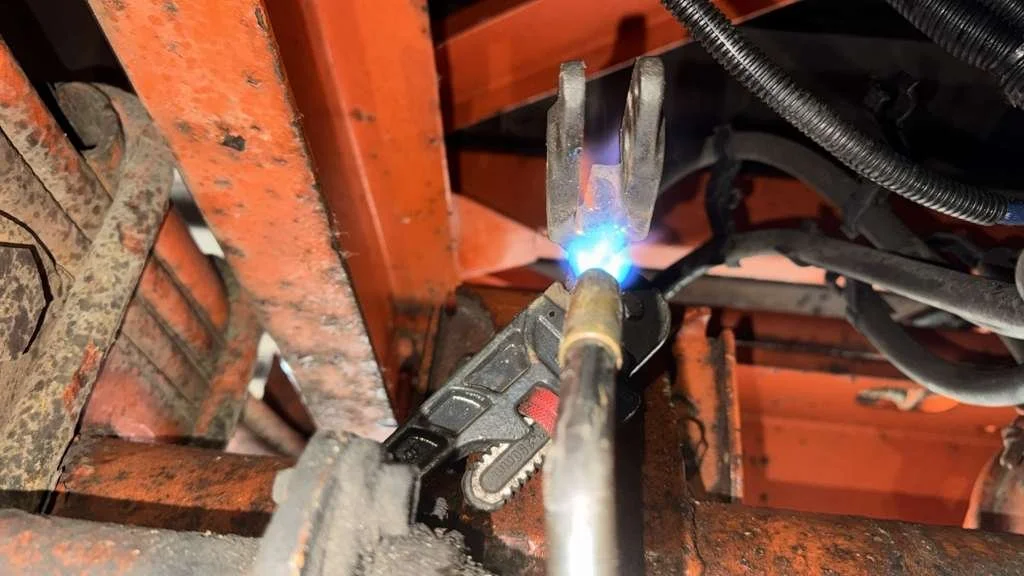 Welding a metal pipe or rod in a workshop setting. The welding tool is situated under a wooden orange-painted structure with visible wires and rusty metal parts around.