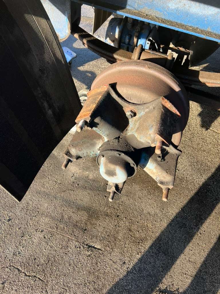 A rusty vehicle wheel hub with five wheel studs attached, missing the wheel, on a paved surface. Part of the vehicle's suspension and brake system are visible behind it.