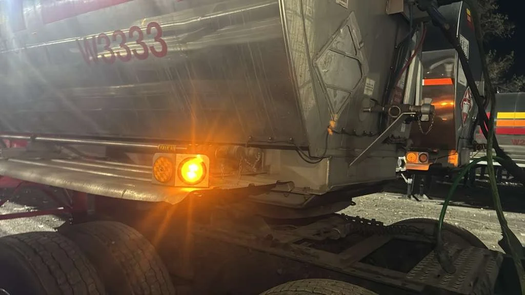 Close-up of a large train car or tanker with an orange light on, parked on railway tracks, with hoses connected to it and other equipment around.