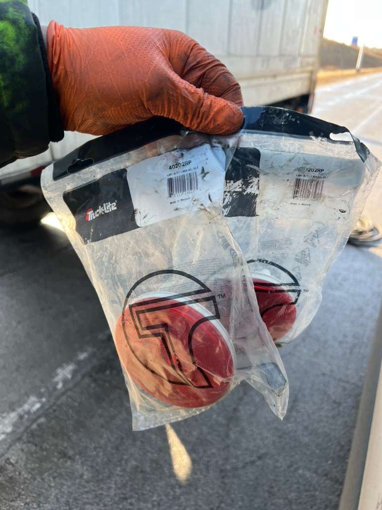 Person wearing orange gloves holding a plastic bag with two red apples inside, near a truck on a paved road.
