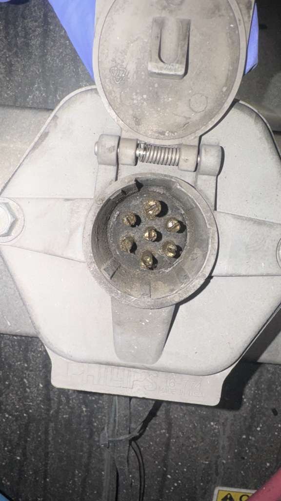 A close-up of an industrial or commercial power outlet with a protective cover, showing six holes for electrical prongs, surrounded by dust or debris.