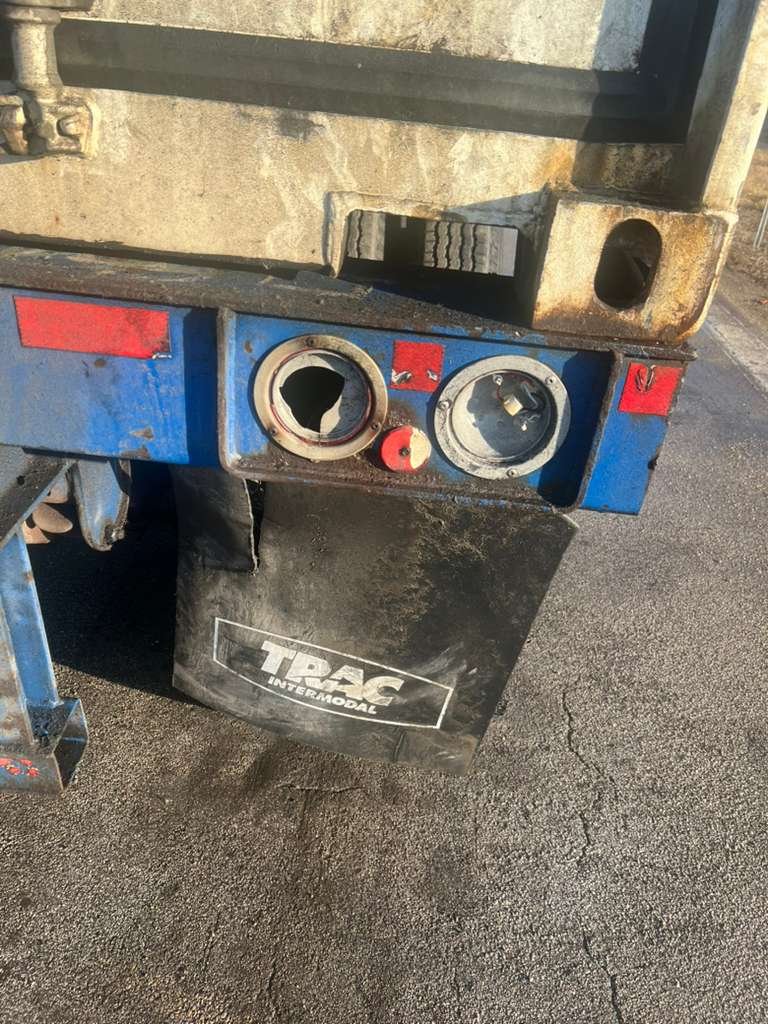 Close-up of a blue and black industrial equipment panel with two circular openings, one damaged. The panel is mounted on a metal frame with a black tarp or cover labeled 'TBA INTERMODAL'. The background shows asphalt pavement with cracks.