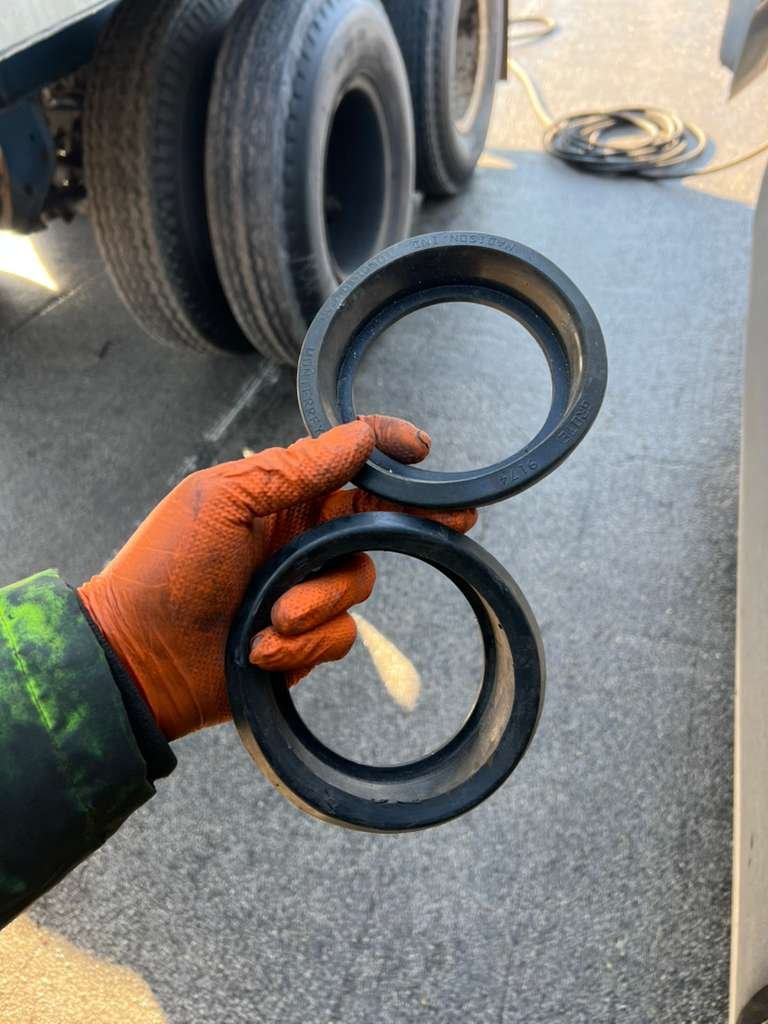 A person wearing an orange glove holding two large black rubber seals in front of a truck's wheels, on a gray asphalt surface.
