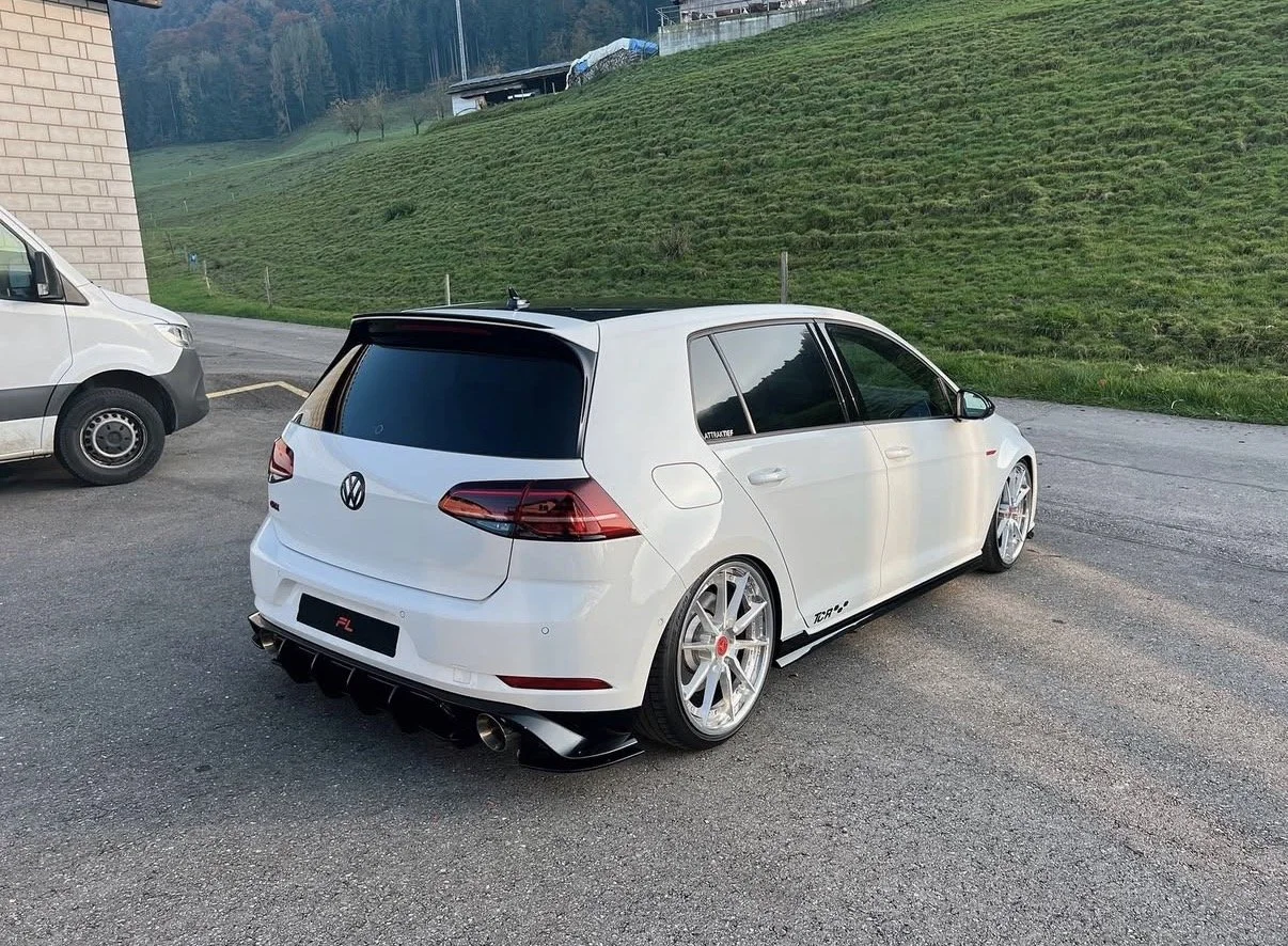 Weißer Volkswagen Golf mit sportlichem Bodykit, großen Rädern und dunklen Scheiben, parked auf einem Parkplatz vor einer grünen, hügeligen Landschaft.