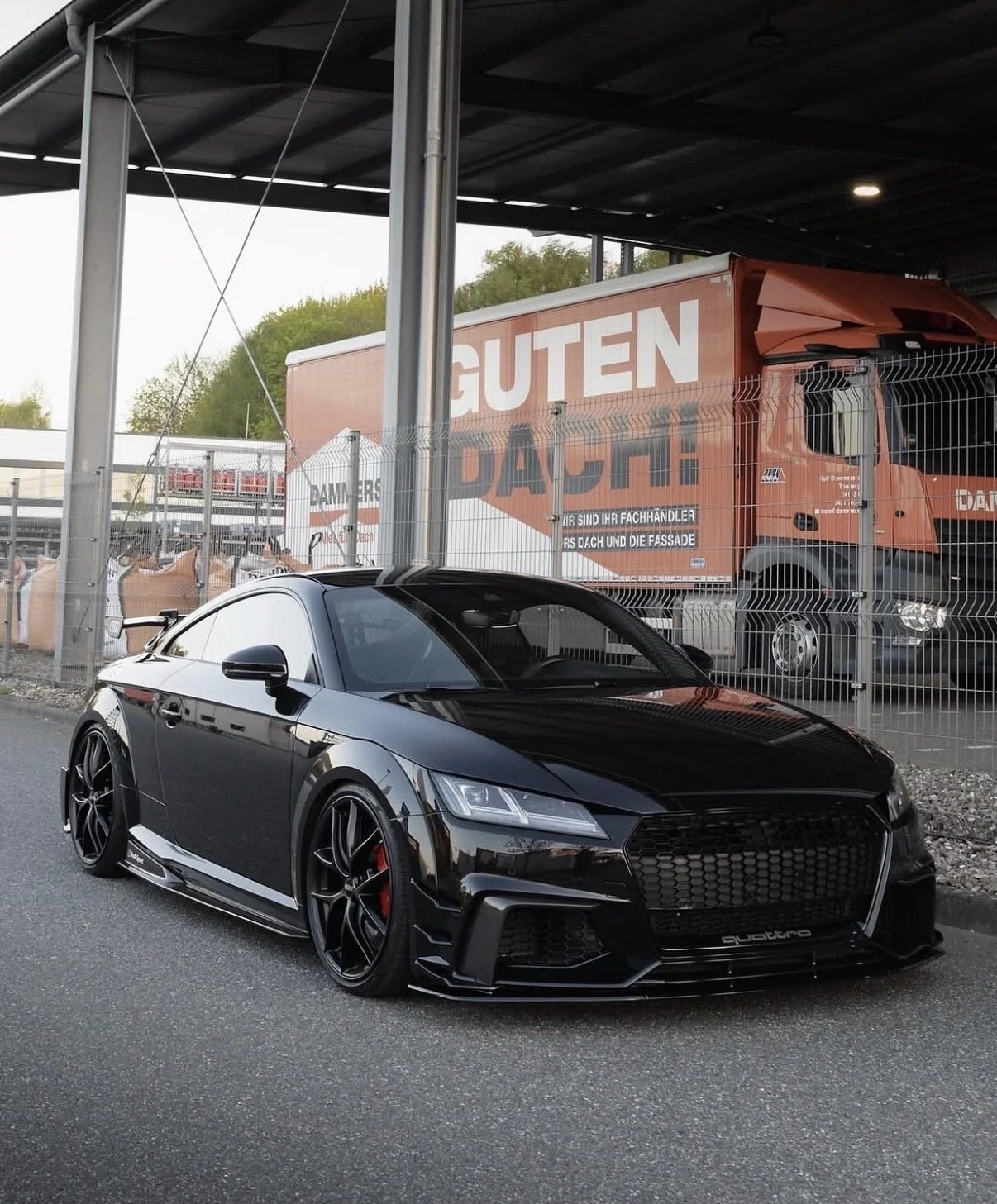 Ein schwarzes Sportauto, vermutlich ein Audi, steht auf einer Straße, im Hintergrund ein Lager mit einem LKW, der Hello-Guten-Dach-Werbung trägt.