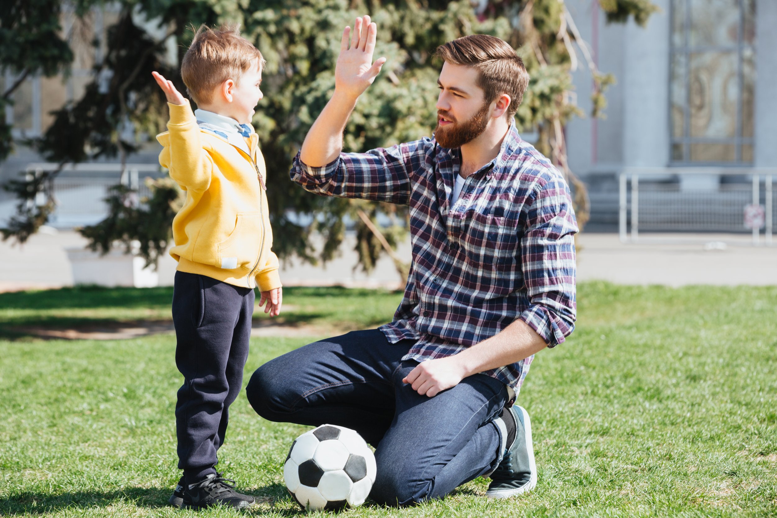 A man kneeling on grass giving a high five to a young boy standing in front of him. There is a soccer ball on the ground between them. The scene appears to be at a park or open field with trees and buildings in the background, and both are smiling.