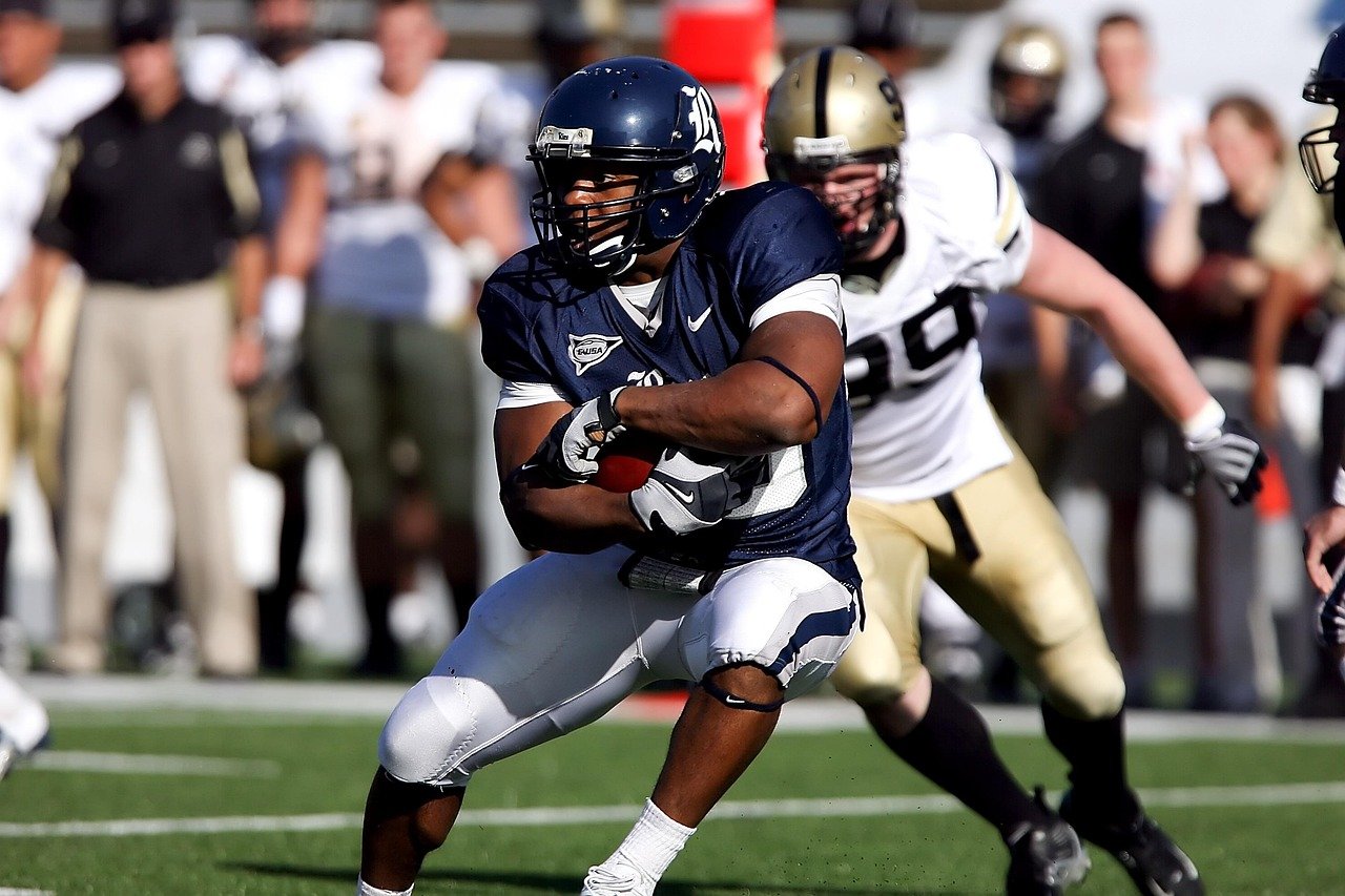 An American football player in a navy blue uniform and helmet carrying the ball, closely pursued by a player in a white and gold uniform on a football field with spectators and coaches in the background.