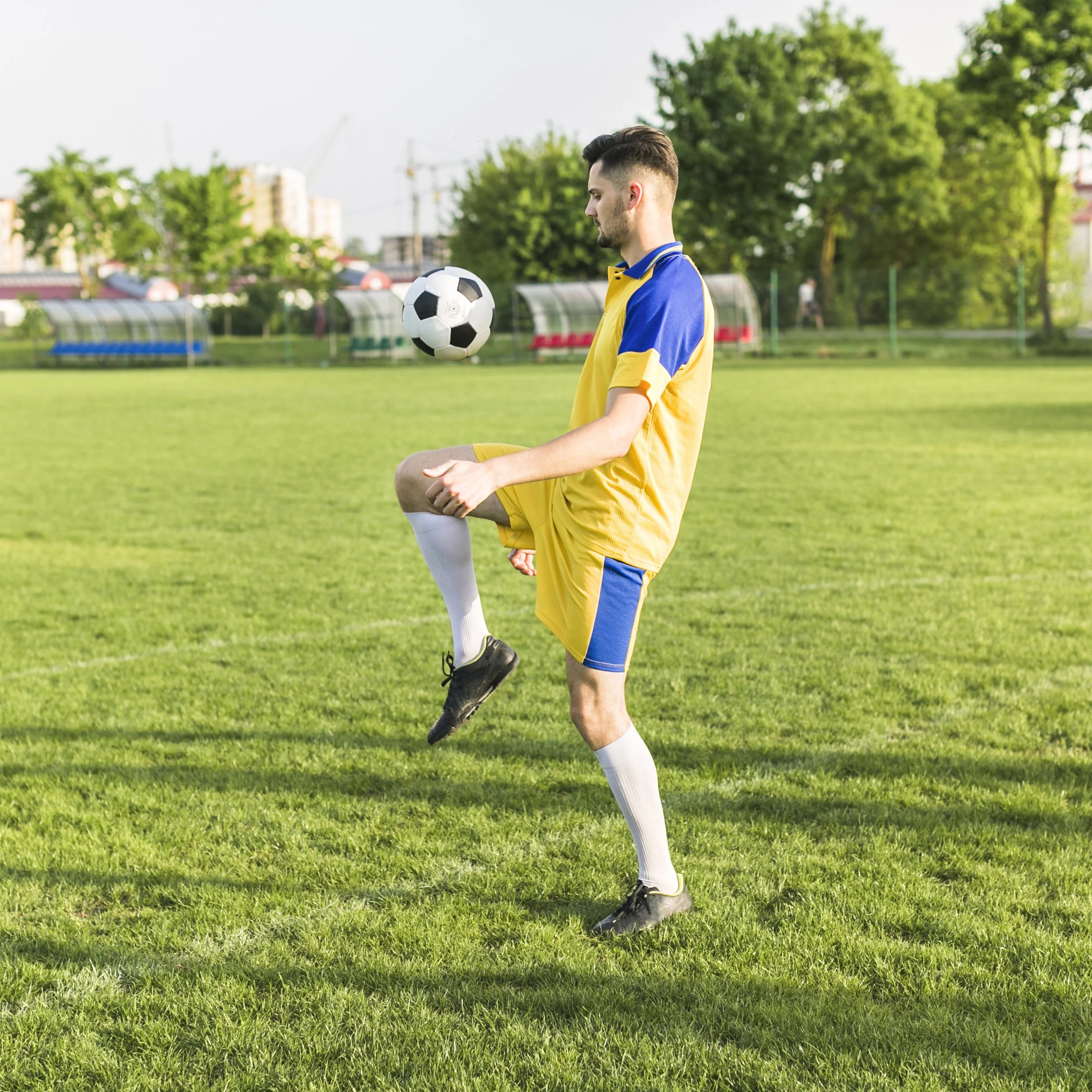 A young male soccer player wearing a yellow and blue uniform practicing juggling a soccer ball on a grassy field with trees and buildings in the background.