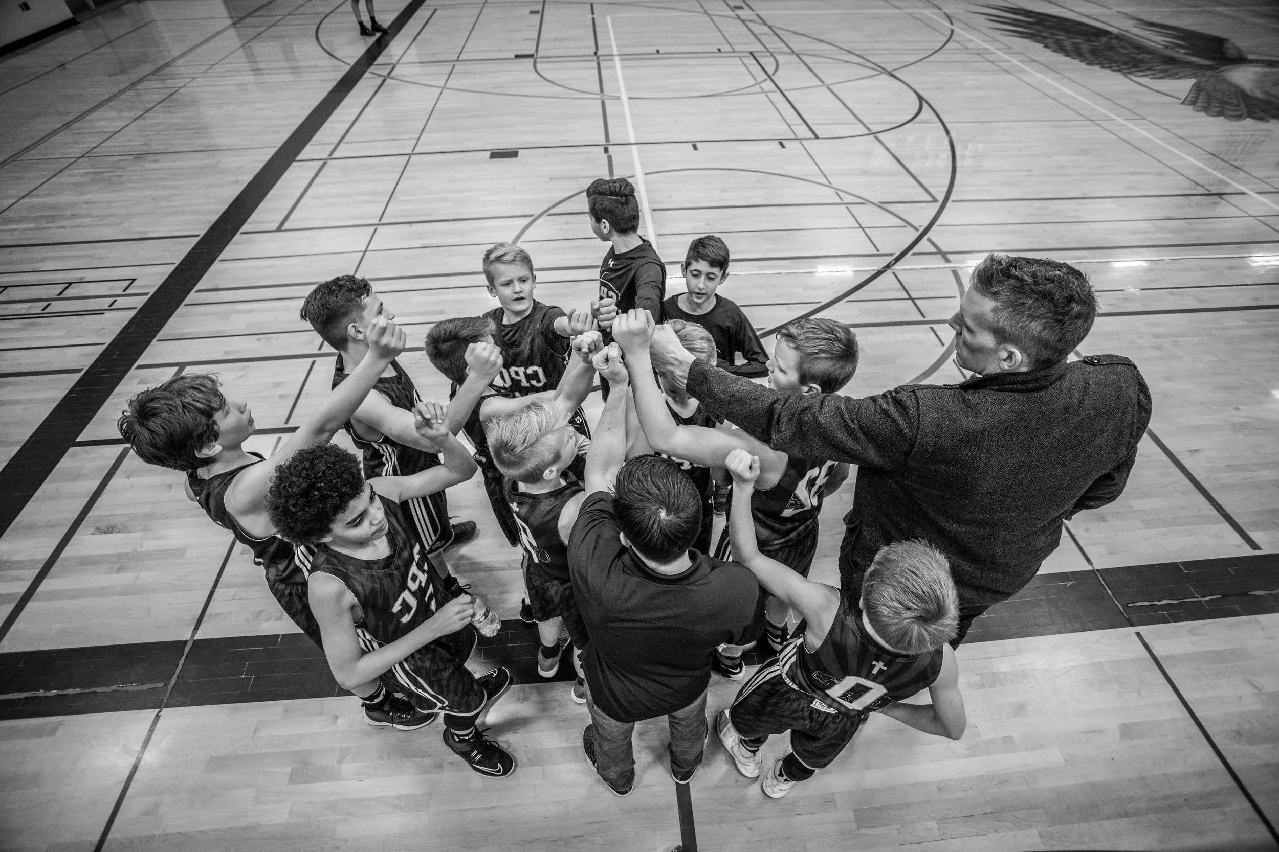 A basketball team of young boys and their coaches celebrating with a huddle on the court.