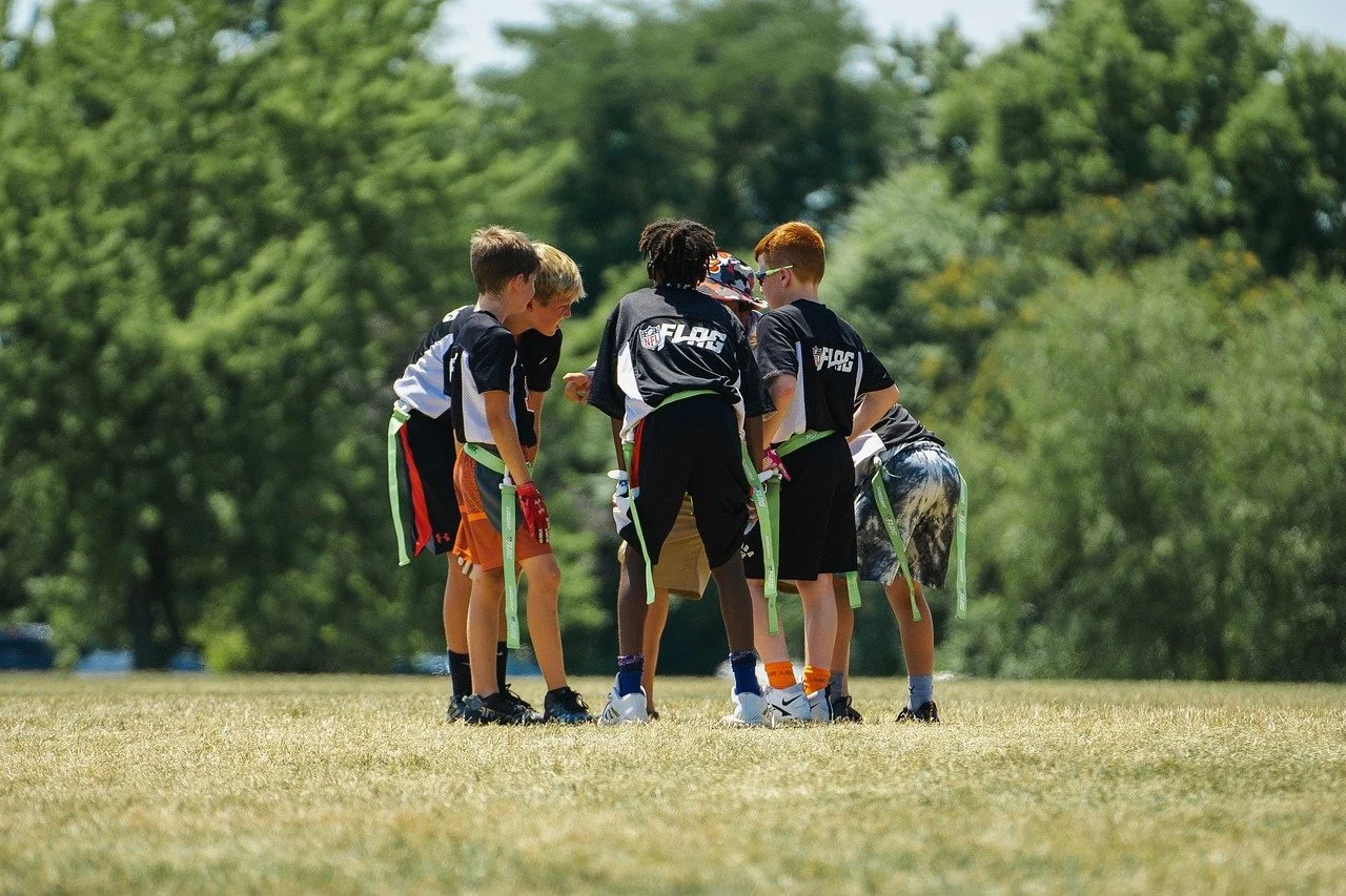 Young boys in football uniforms gathered in a huddle on a grassy field during daytime.