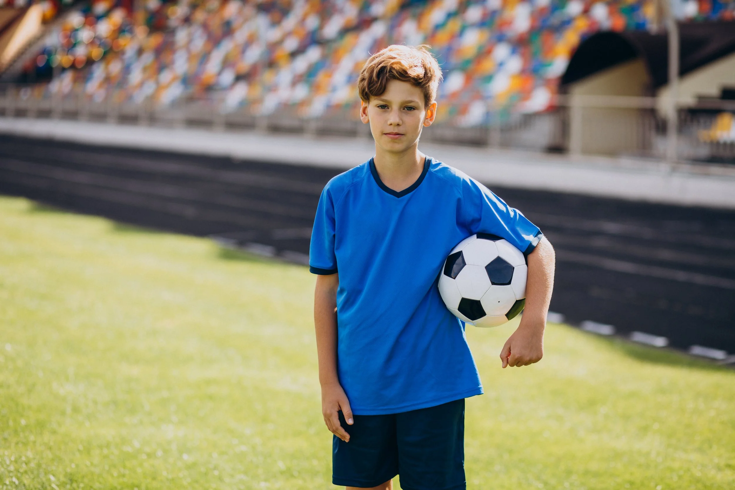 A young boy wearing a blue sports jersey holding a black and white soccer ball on a grass field at a sports stadium.