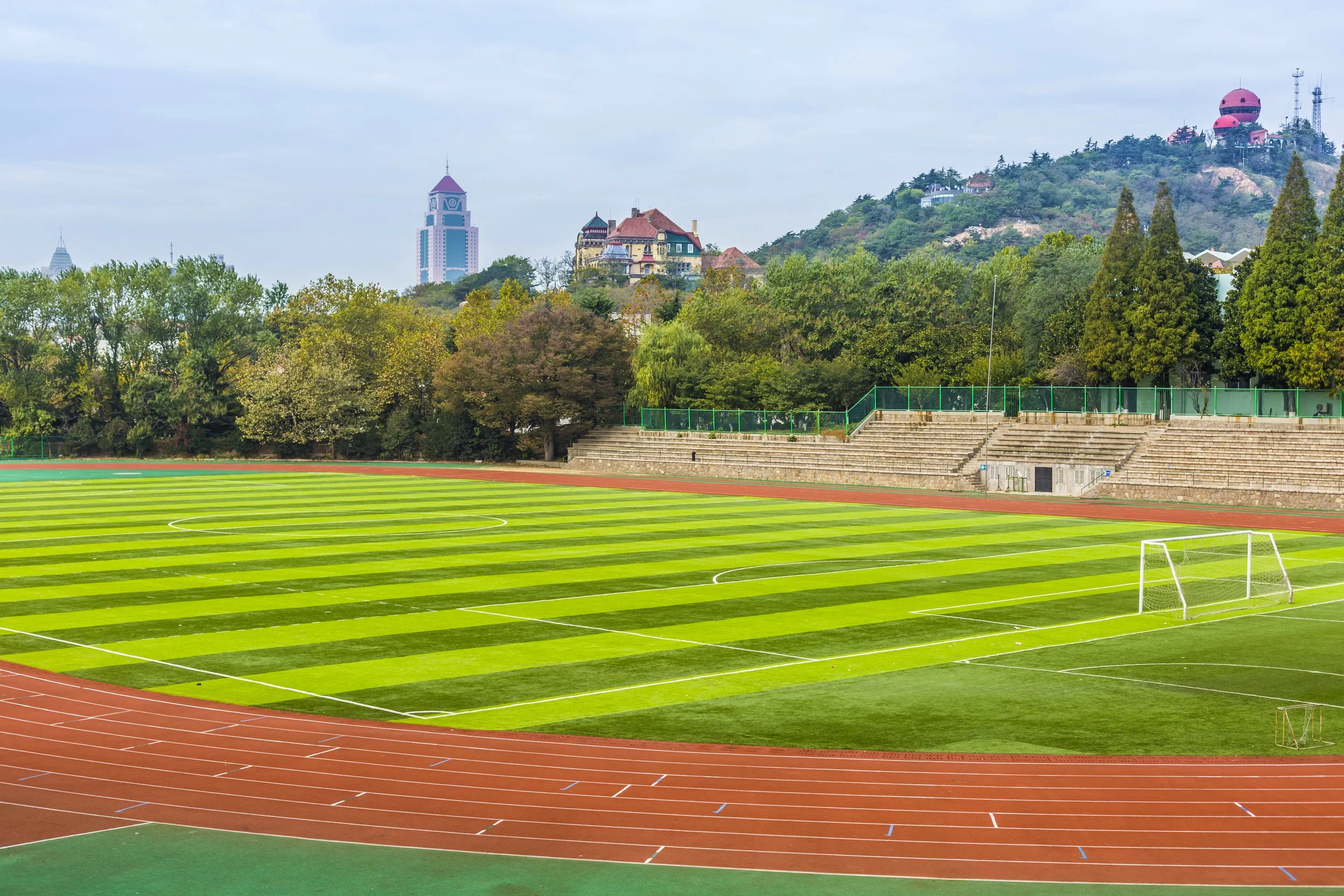 Empty outdoor soccer field with lush green grass, surrounded by a running track, and trees in the background with hills and buildings, including a tower and observatory domes, under a cloudy sky.
