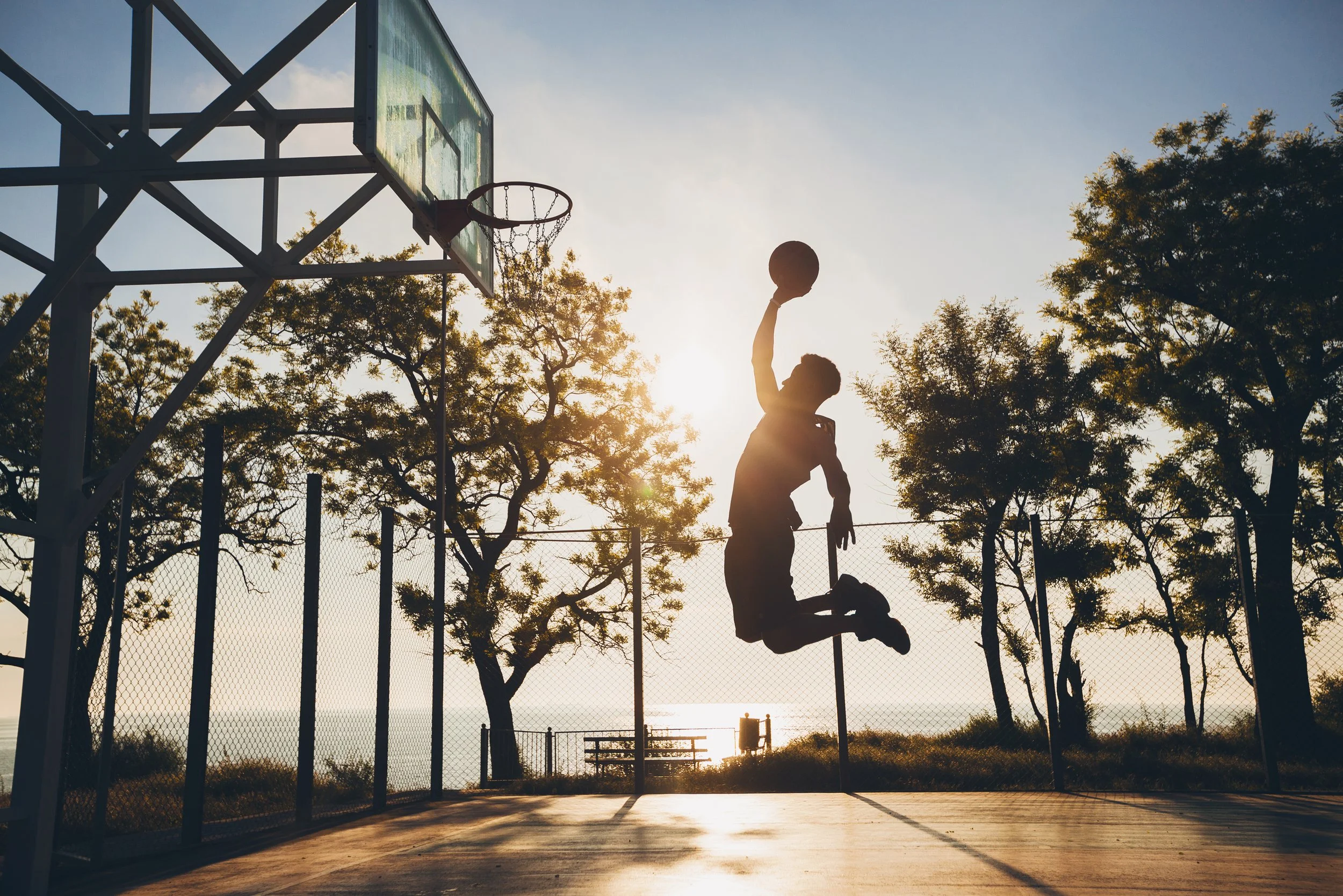Silhouette of a boy jumping to dunk a basketball on an outdoor court during sunset, with trees and a fence in the background.
