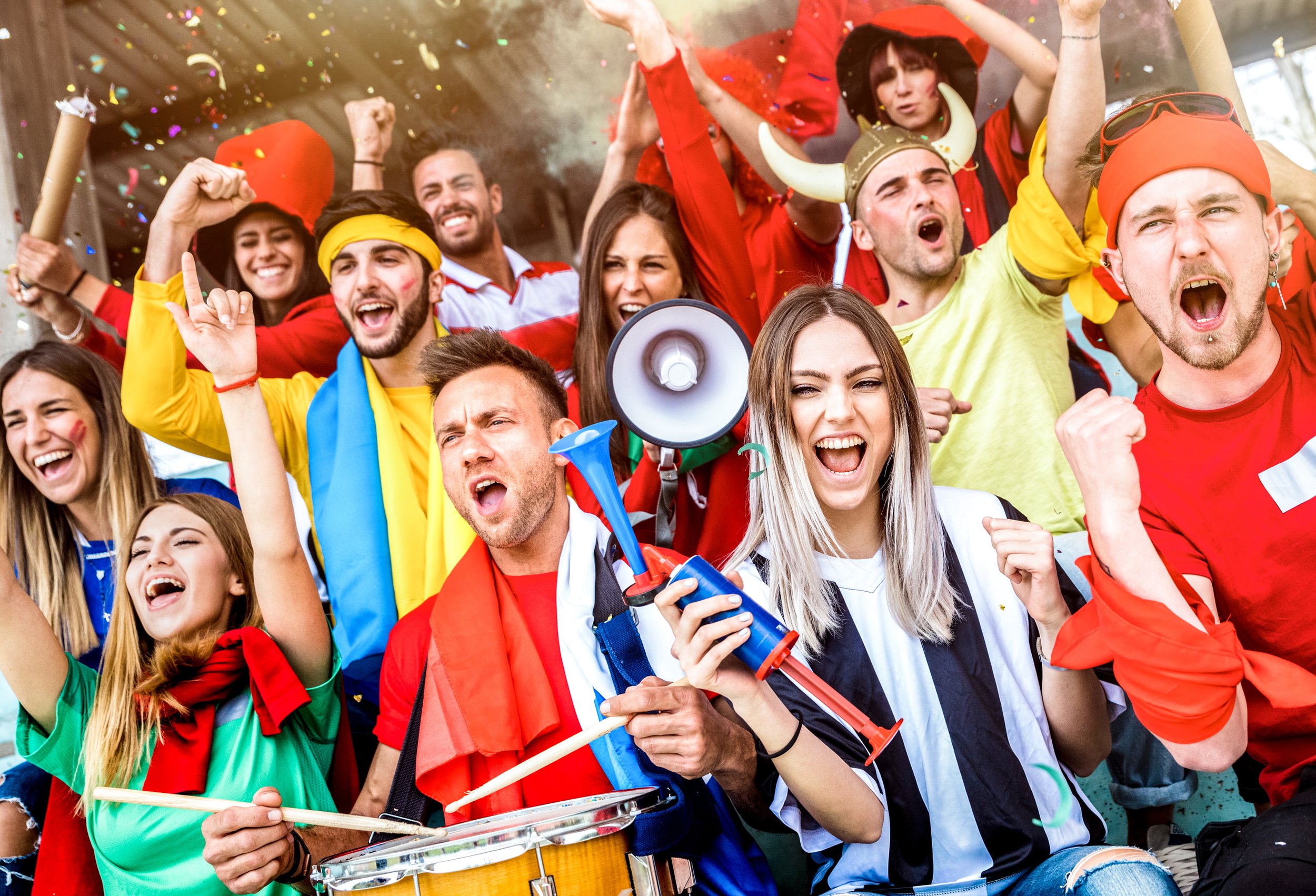 A diverse group of sports fans celebrating passionately in a stadium, wearing colorful clothing and accessories, with confetti in the air and smiling faces.