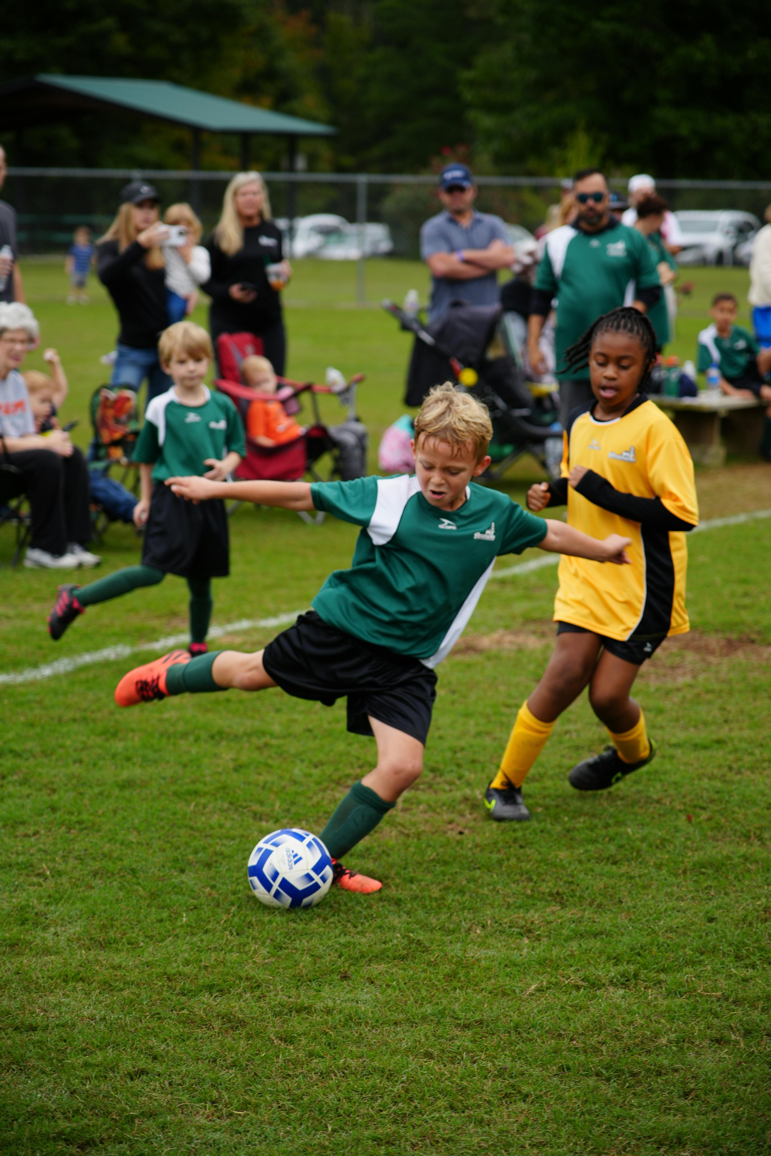 Children playing soccer on a grass field, with spectators watching in the background.