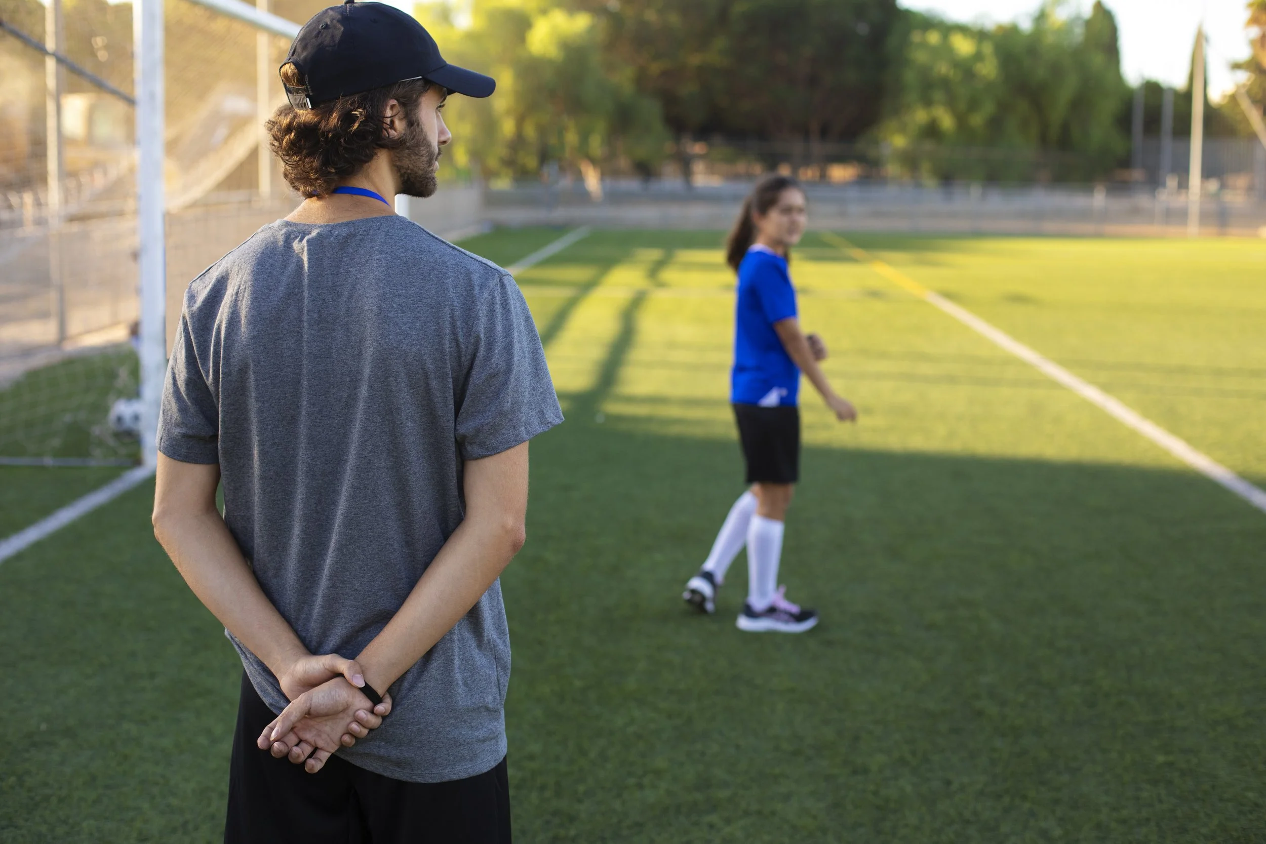 A young man with curly hair, wearing a black cap, gray t-shirt, and black shorts, watching a girl in a blue sports uniform on a soccer field during late afternoon or early evening.