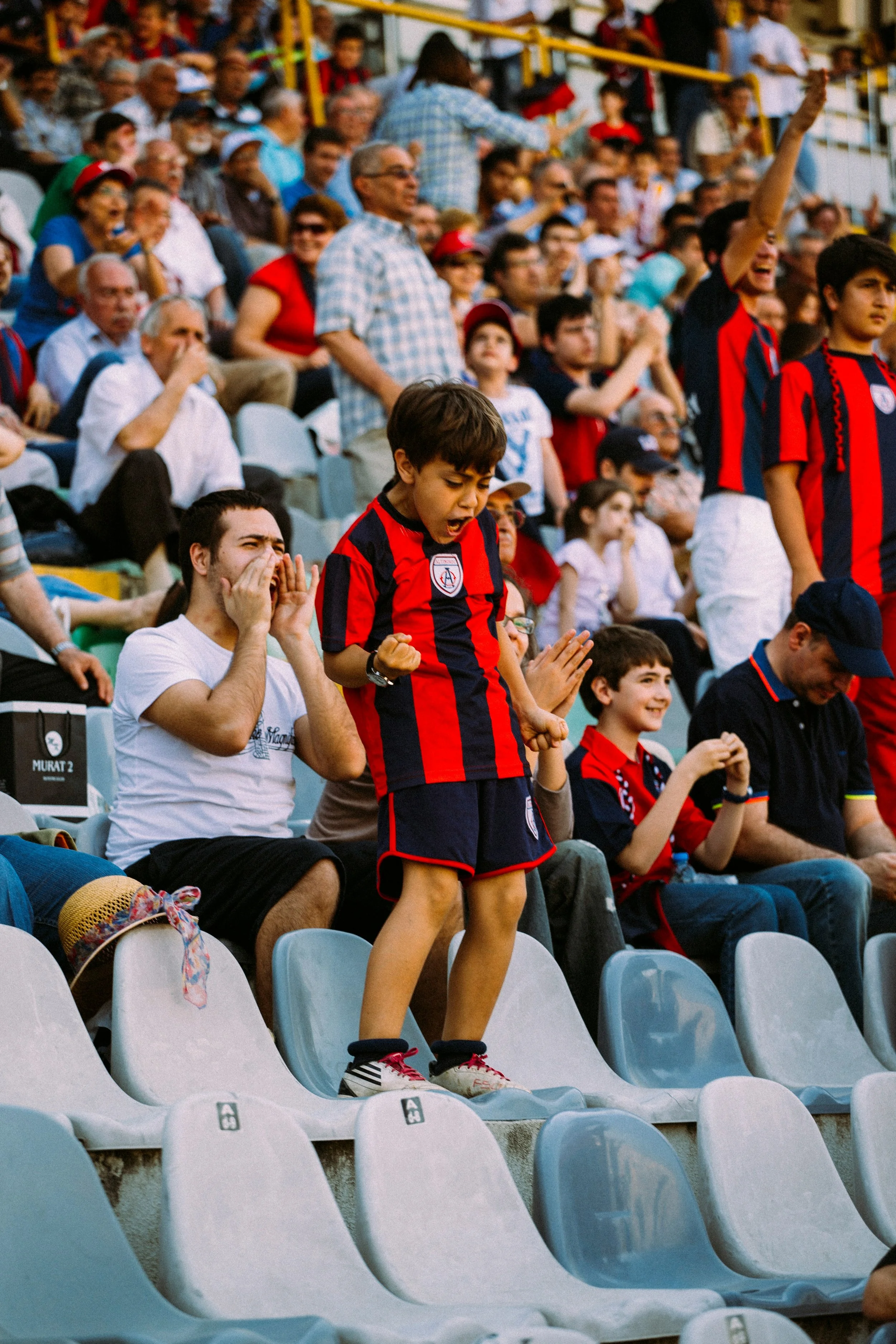 Children and adults in a stadium watching a soccer match, some standing and cheering, many dressed in team jerseys and casual clothing.