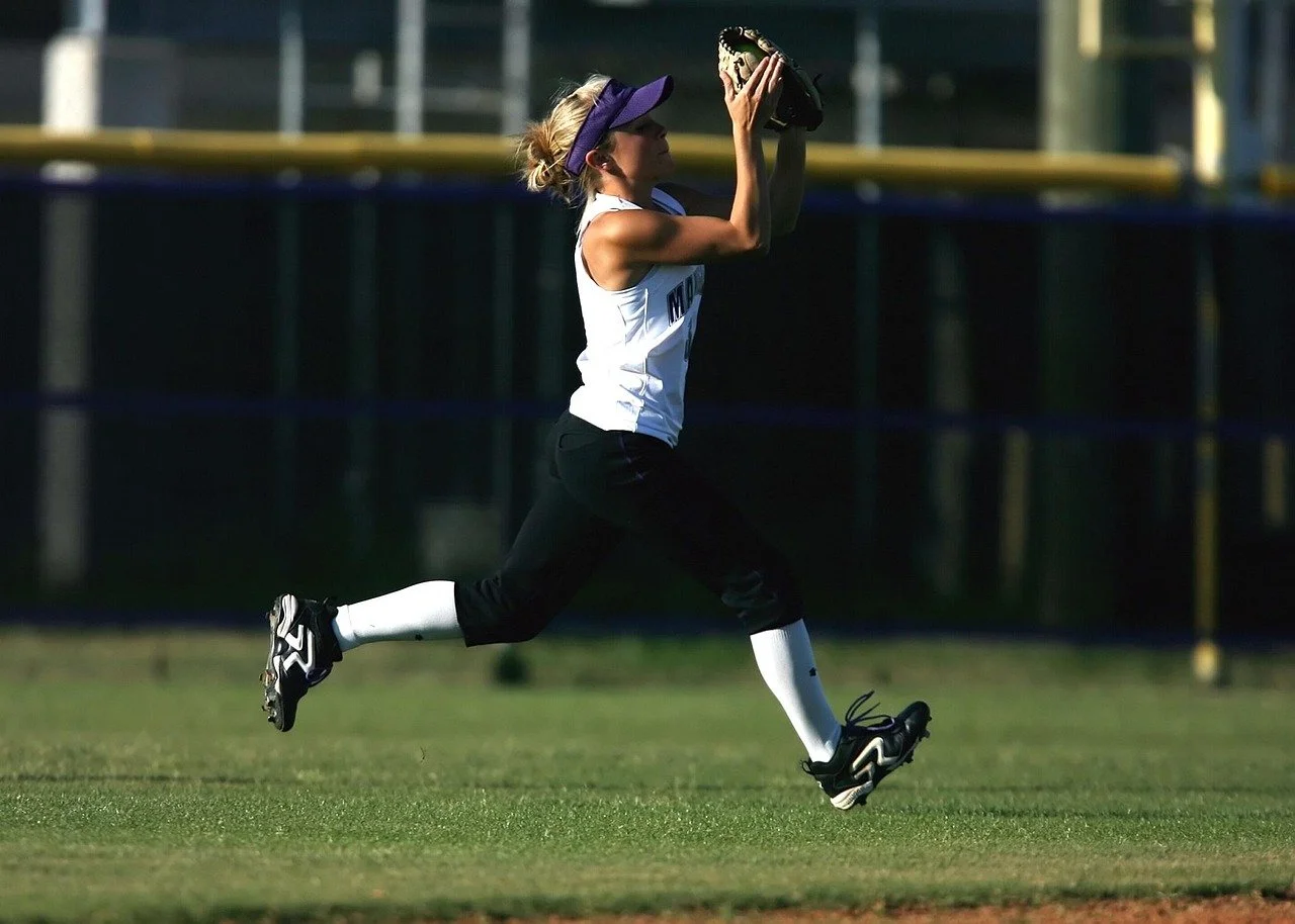 A woman playing softball on a grassy field, wearing a white sports jersey, black pants, white knee-high socks, and black cleats, with a purple visor cap, catching a ball with her glove.