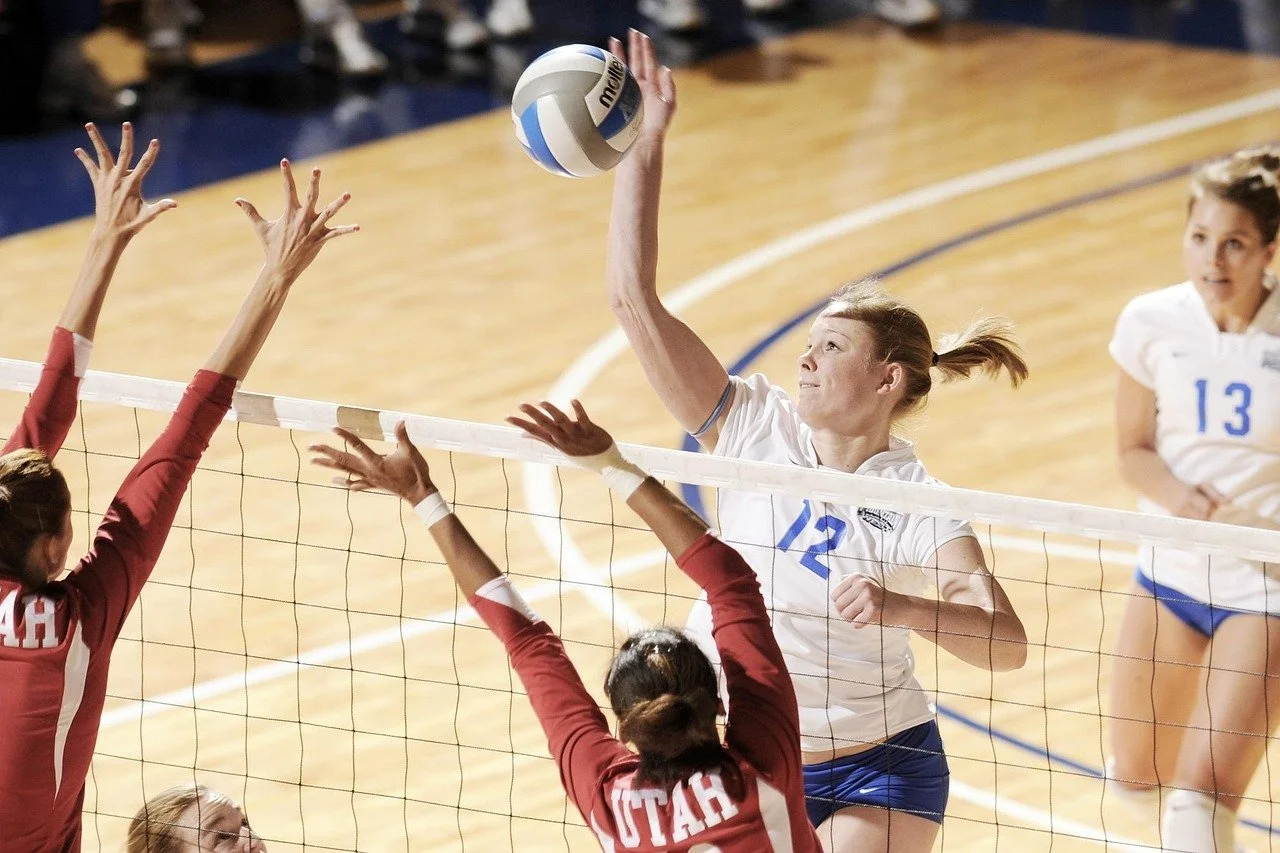 A female volleyball player from the team with a white jersey numbered 12 is spiking the volleyball over the net while players from the opposing team with red jerseys are jumping to block.