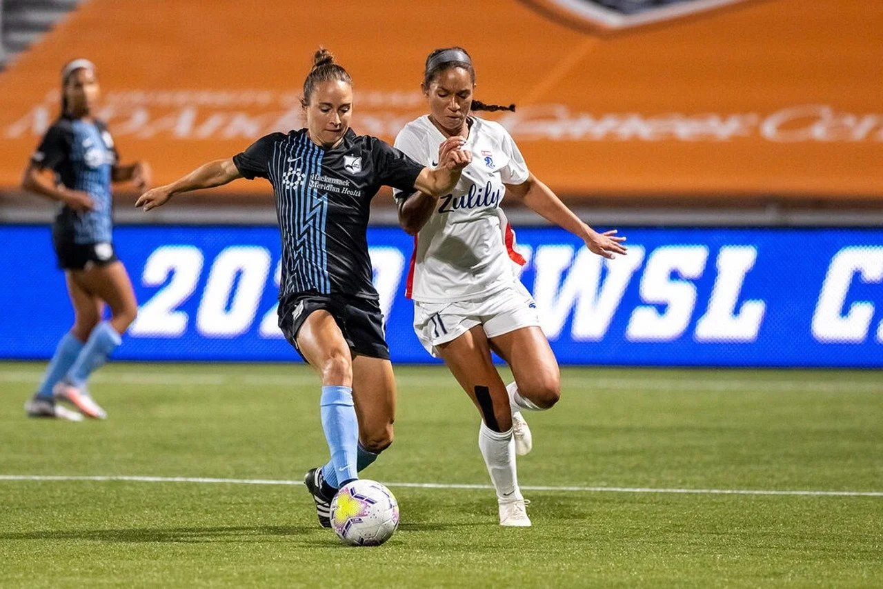 Two female soccer players competing for control of the ball on a soccer field, with another player in the background, during a match.