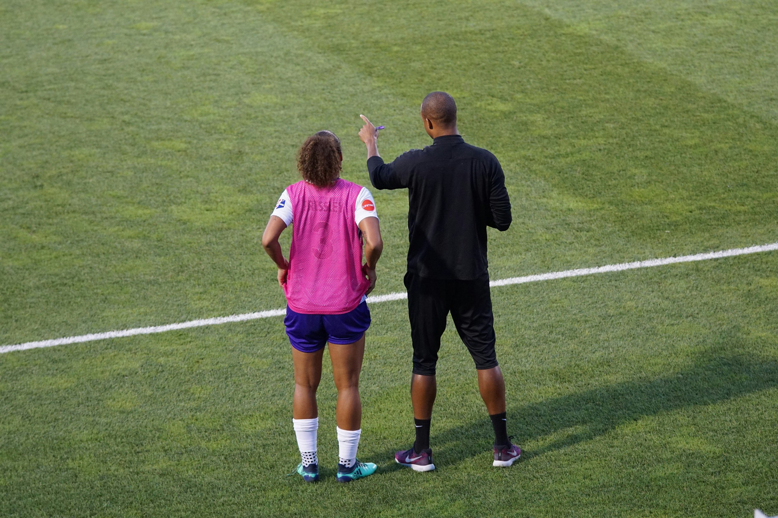 A soccer coach and a player talking on the field. The player is wearing a pink vest over a white jersey, blue shorts, and soccer cleats. The coach is wearing black athletic wear and shoes. The coach appears to be giving instructions or a pep talk.