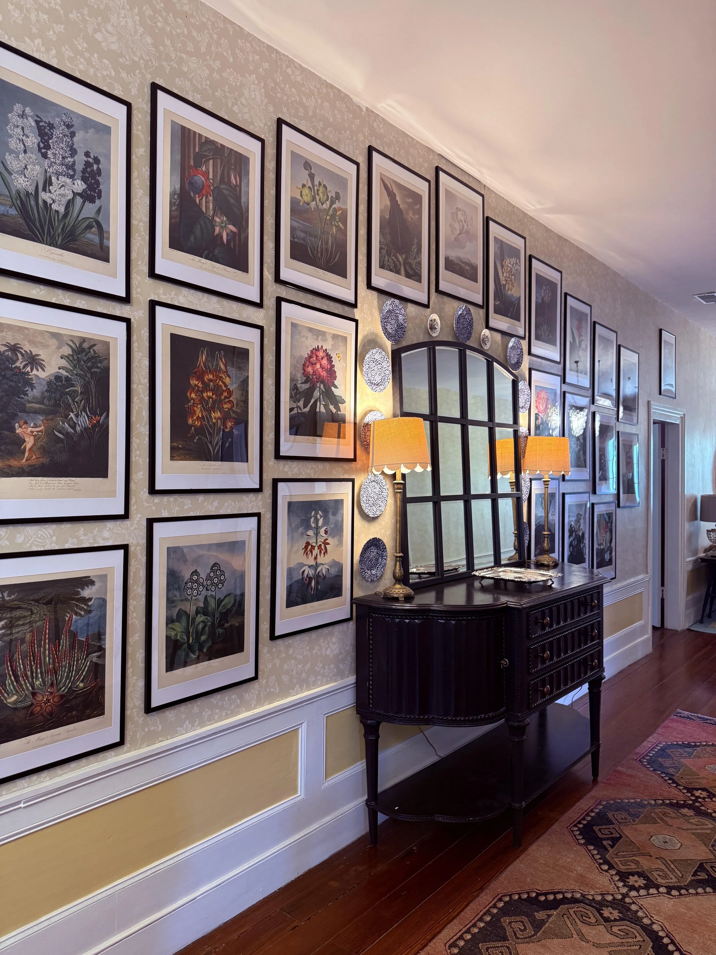 A hallway with a gallery wall of framed botanical illustrations, including various flowers and plants, with two orange table lamps and a black wooden side table, next to a mirror and a decorative plate display.