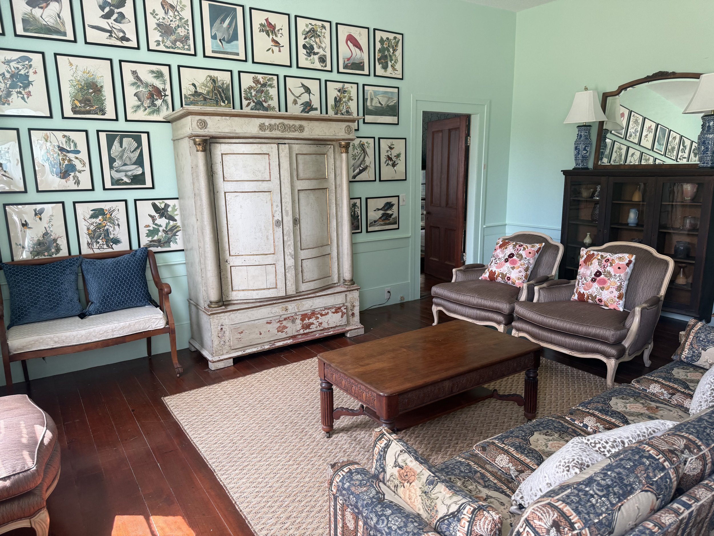 Living room with light green walls decorated with multiple framed artwork of birds, a wooden door, a white vintage fireplace mantel, a dark wood cabinet with decorative vases, and various upholstered chairs and sofas with patterned cushions, a wooden coffee table, a patterned area rug, and a large mirror reflecting part of the room.