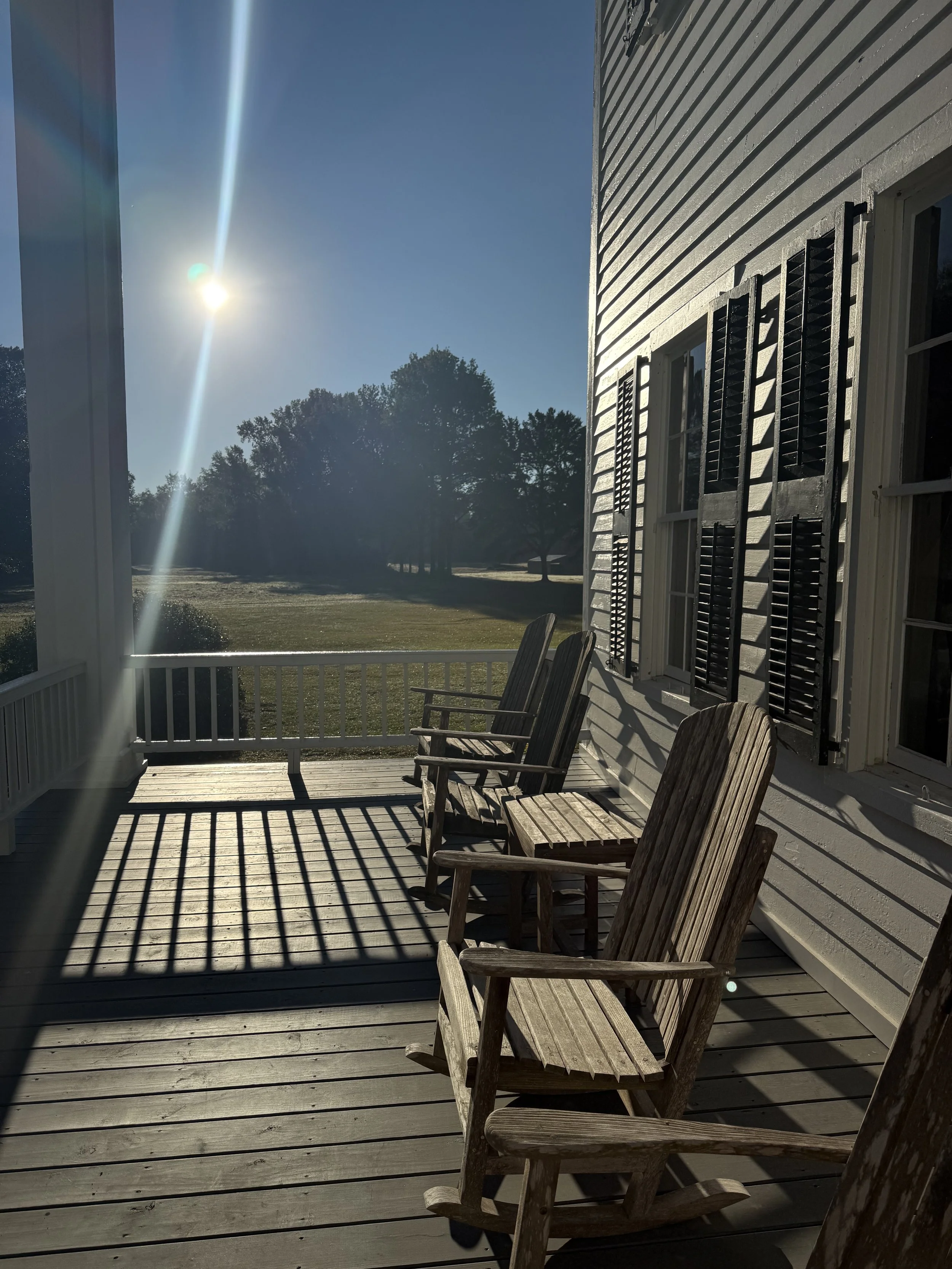 A porch with wooden chairs, shadows cast by the railing, a house with white siding and black shutters, a grassy yard with trees, and the sun shining in a clear blue sky.