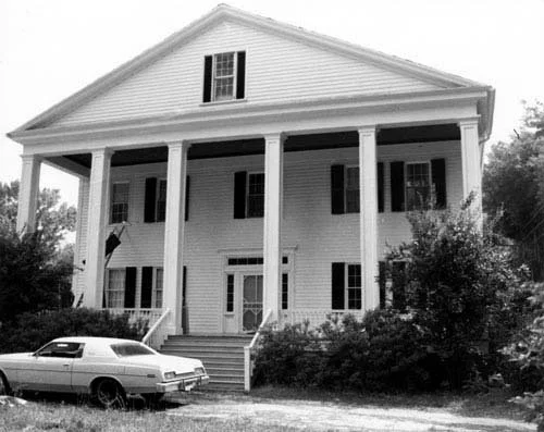 The large, two-story white estate at Rip Raps with a front porch supported by tall columns, black shutters on the windows, and a vintage car parked in front.