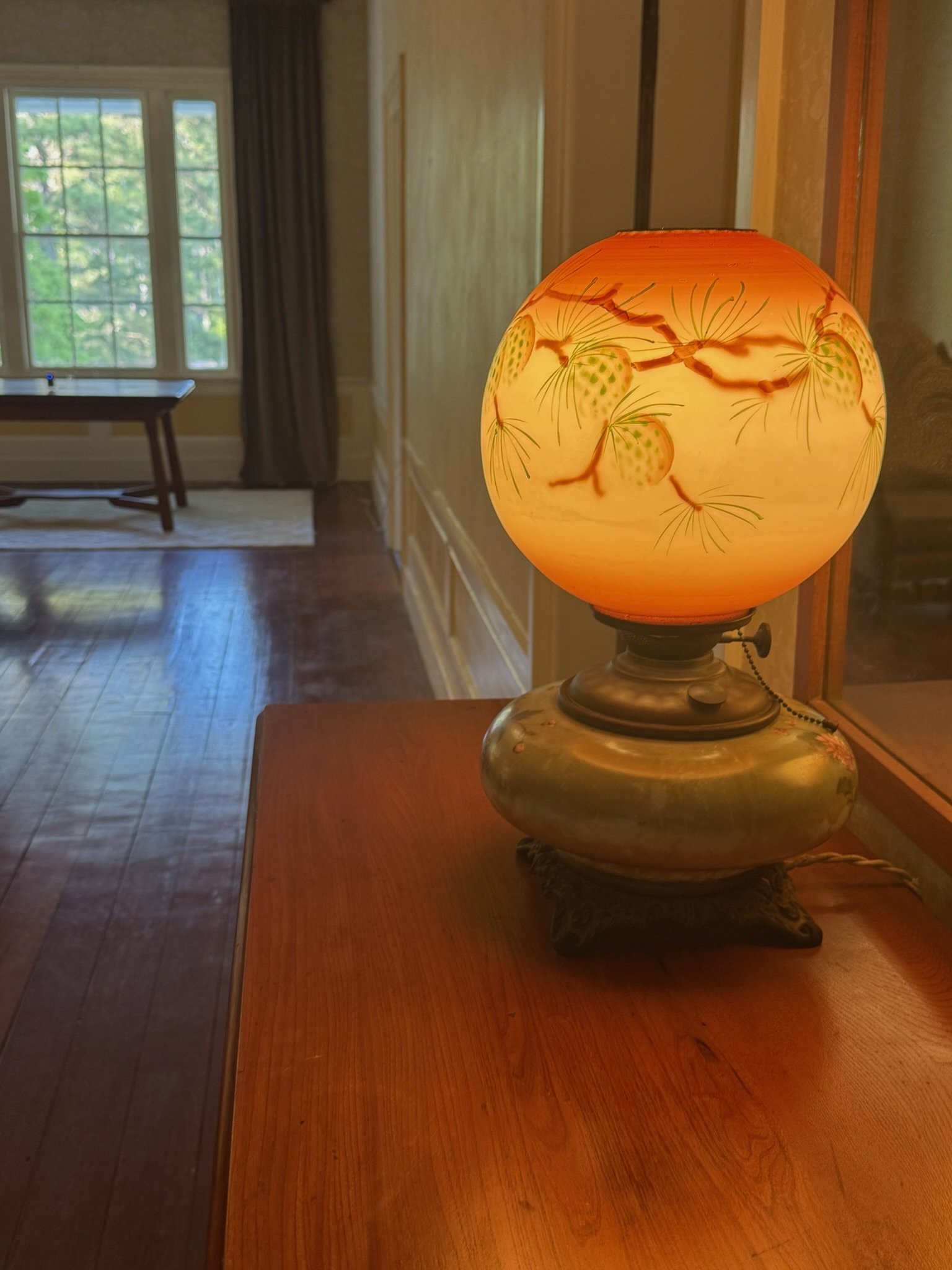 An ornate table lamp with a decorative shade featuring pine cone and pine needle designs, sitting on a wooden table in a warmly lit room with large windows and dark curtains.