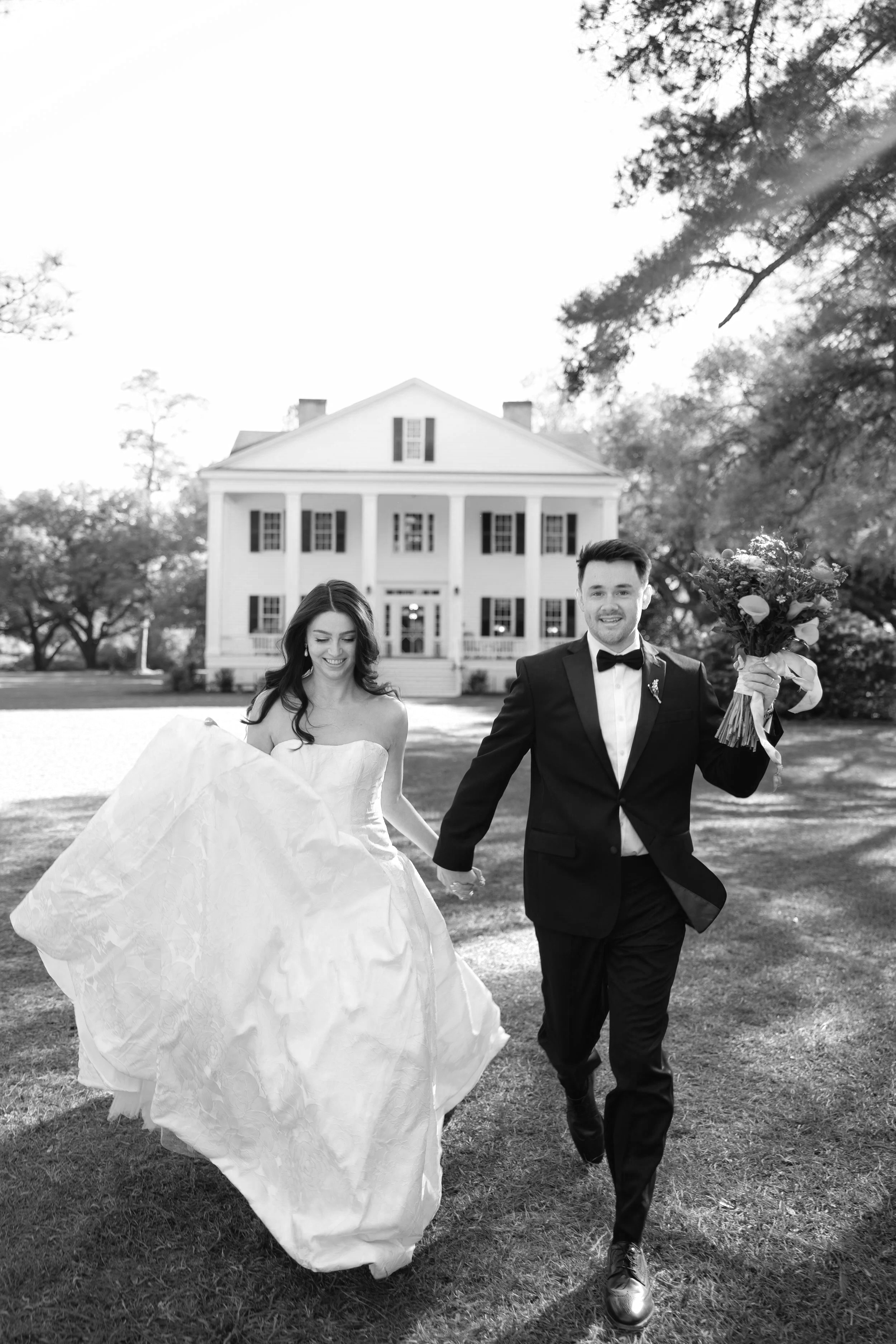 A newlywed couple walking hand in hand on a lawn in front of a large white house, with the bride holding her dress and the groom holding a bouquet of flowers, in black and white at Rip Raps.