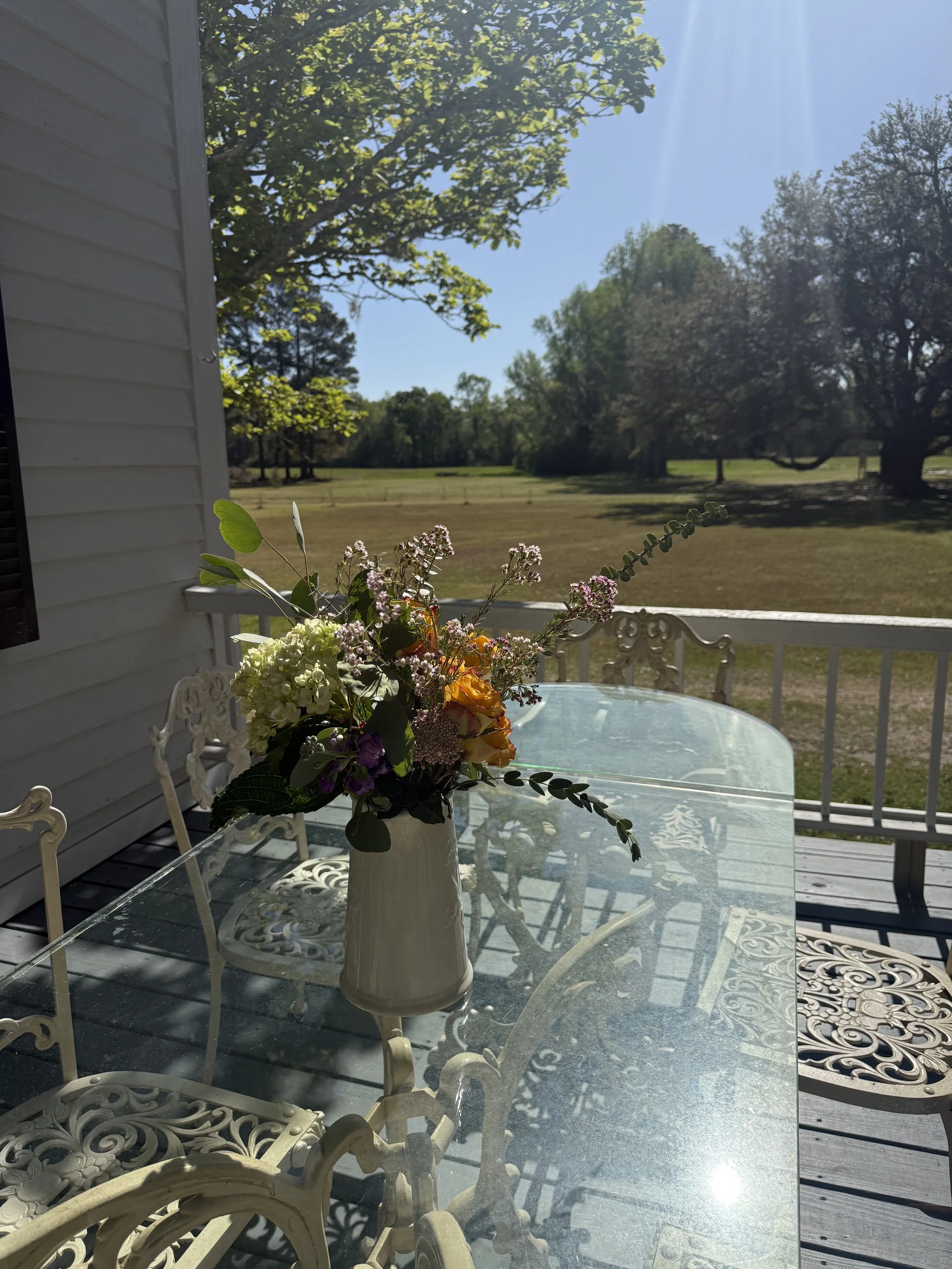 A glass-top outdoor table with a cream-colored vase holding a mixed flower bouquet, on a porch with white ornate metal chairs and railings, overlooking a grassy yard with large trees and a bright blue sky in the background.