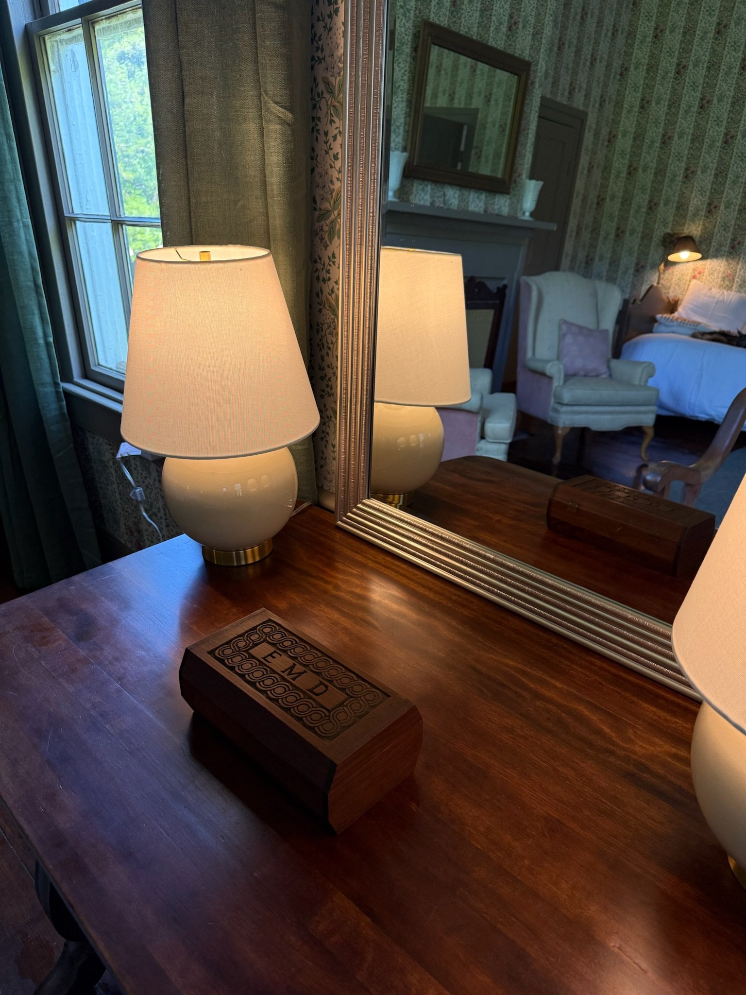 A wooden table with a decorative box and a table lamp, seen in front of a large mirror reflecting a cozy room with vintage furniture, floral wallpaper, and a window with green curtains.
