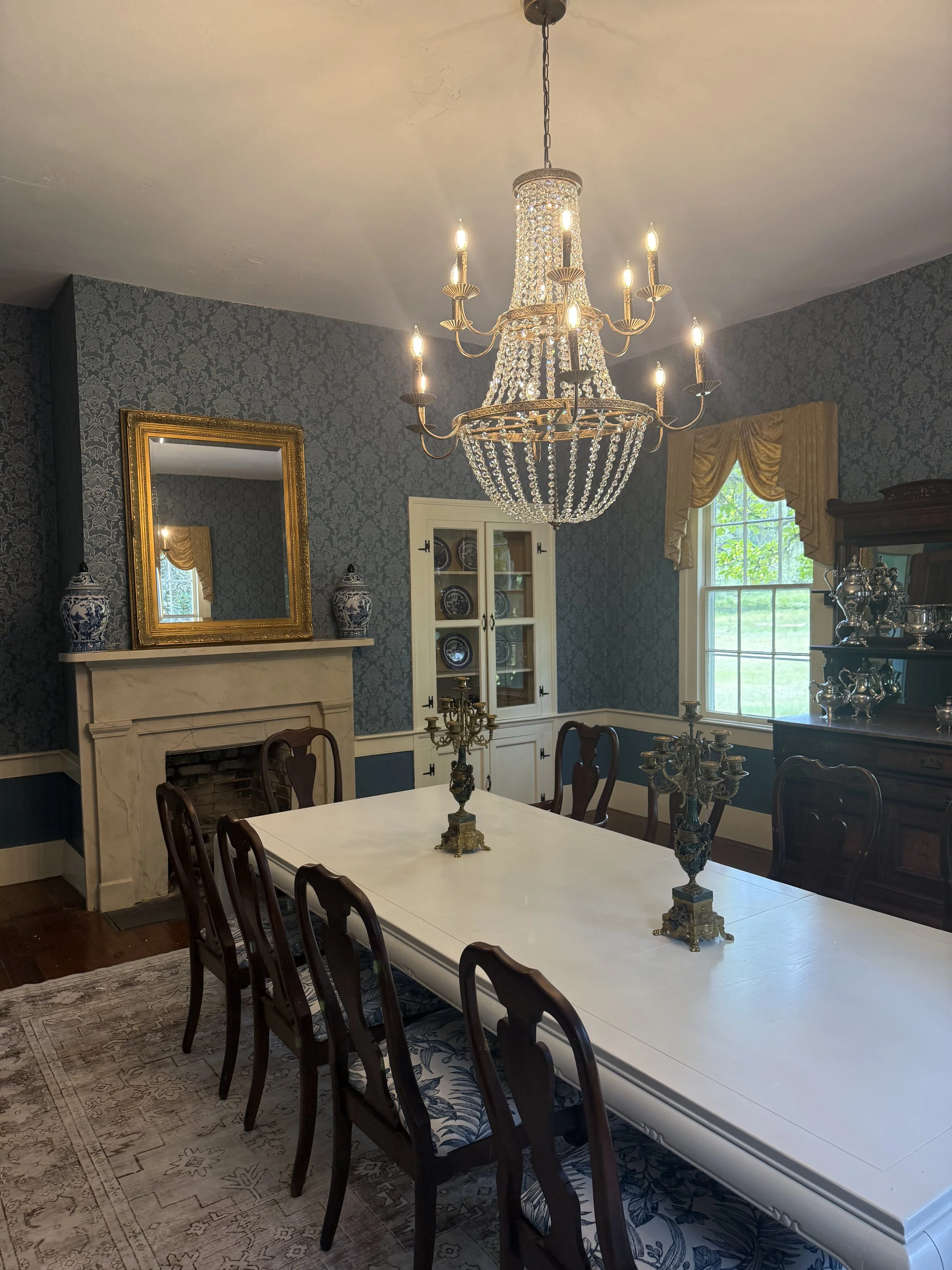 A formal dining room with a long white table and eight dark wooden chairs. The room features a large chandelier with crystal decorations hanging from the ceiling, a marble fireplace with a gold-framed mirror above, and a window with gold curtains showing greenery outside. The walls are covered with dark damask wallpaper.