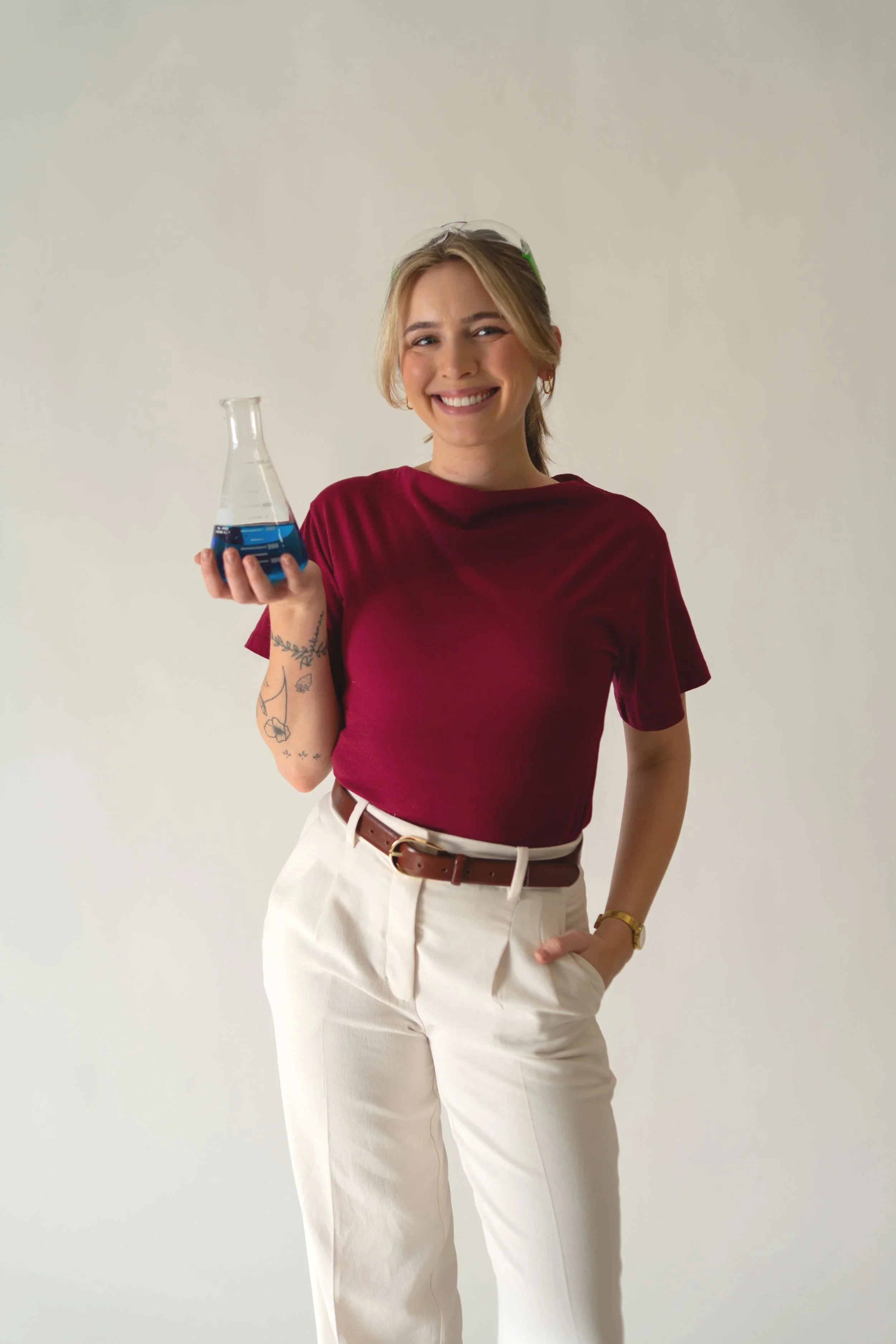 Young woman with tattoos on her left arm, wearing a maroon shirt and white pants, holding a beaker with blue liquid, smiling, standing against a plain light background.