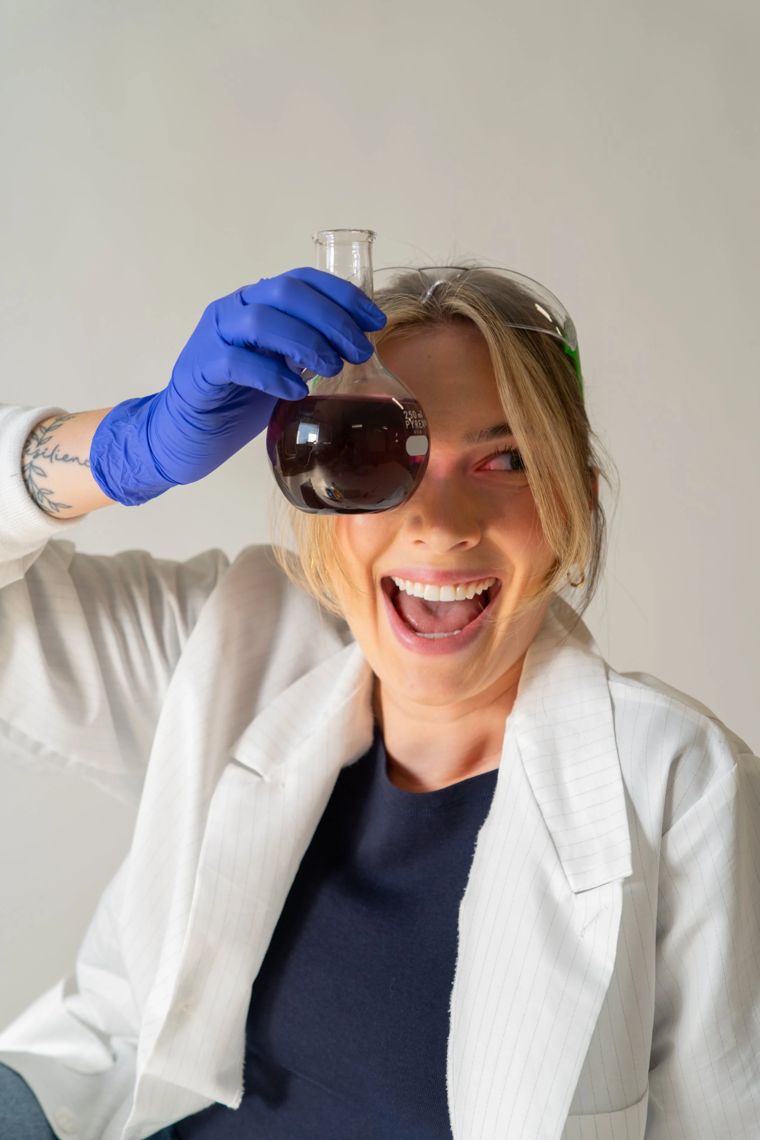 A woman wearing a white lab coat and blue gloves holding a chemistry flask with a dark liquid, smiling and looking playful.