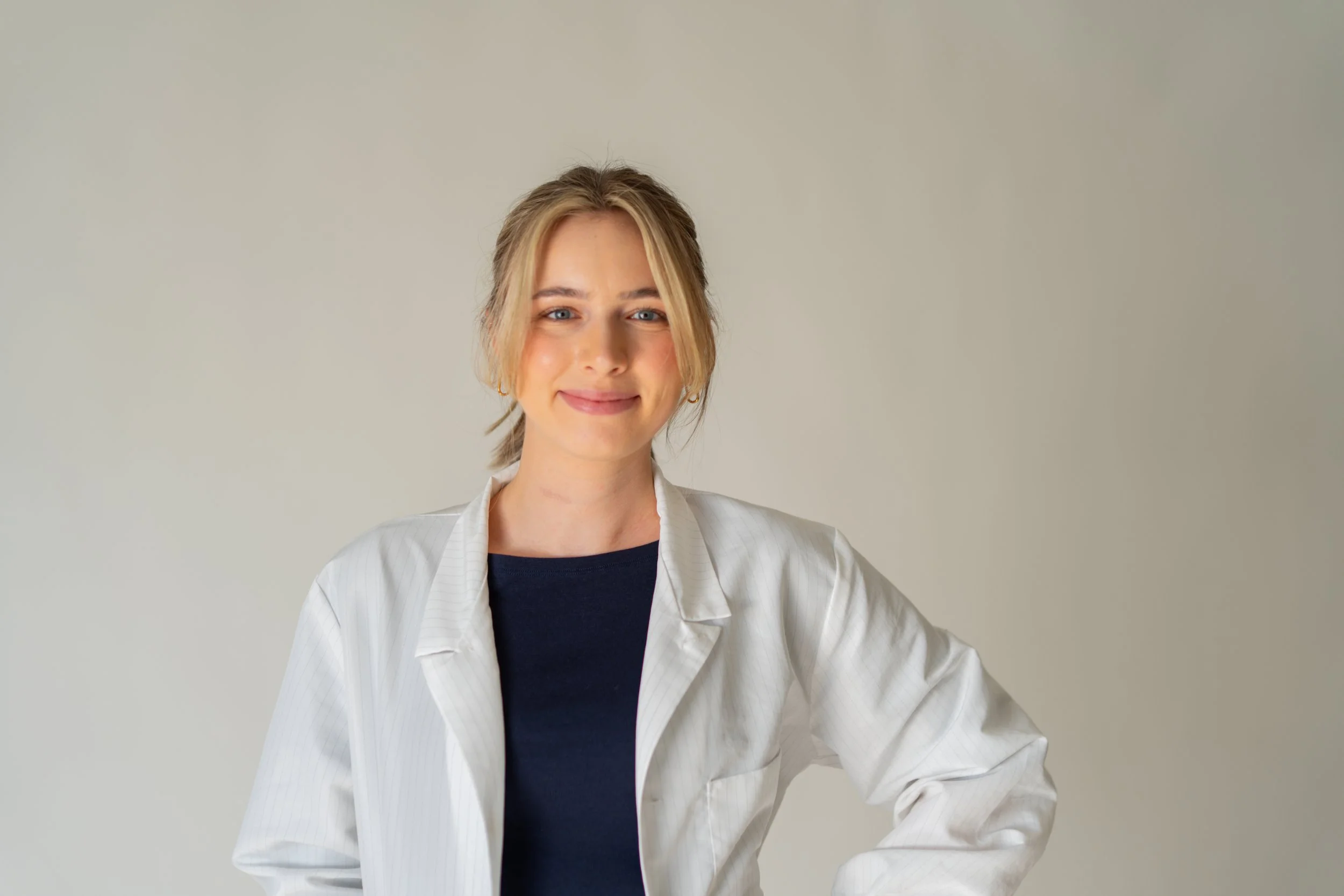 A young woman with blonde hair smiling, wearing a white medical coat over a black top, standing against a neutral background.