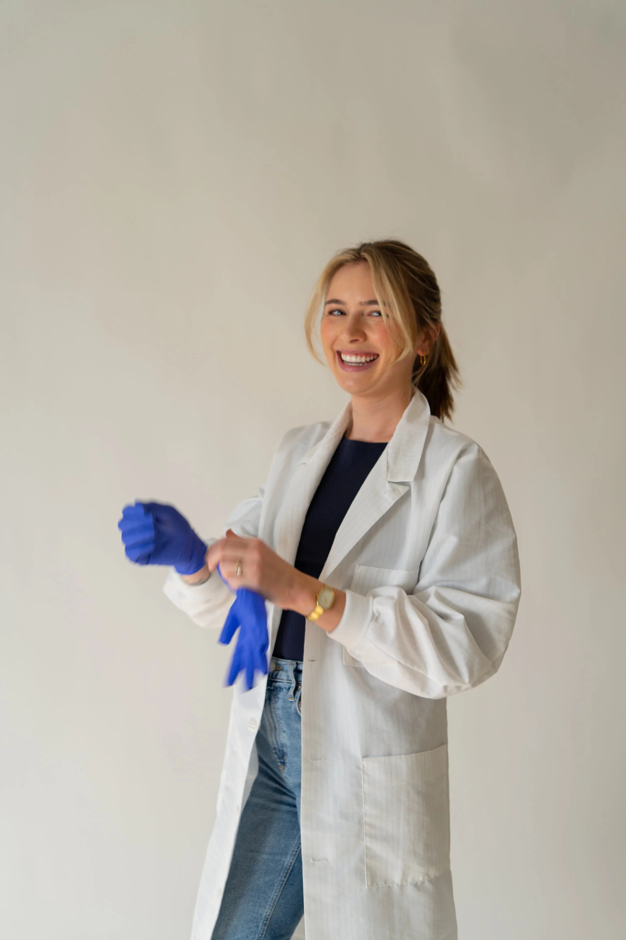 A woman in a white lab coat smiling, holding blue disposable gloves, against a plain background.
