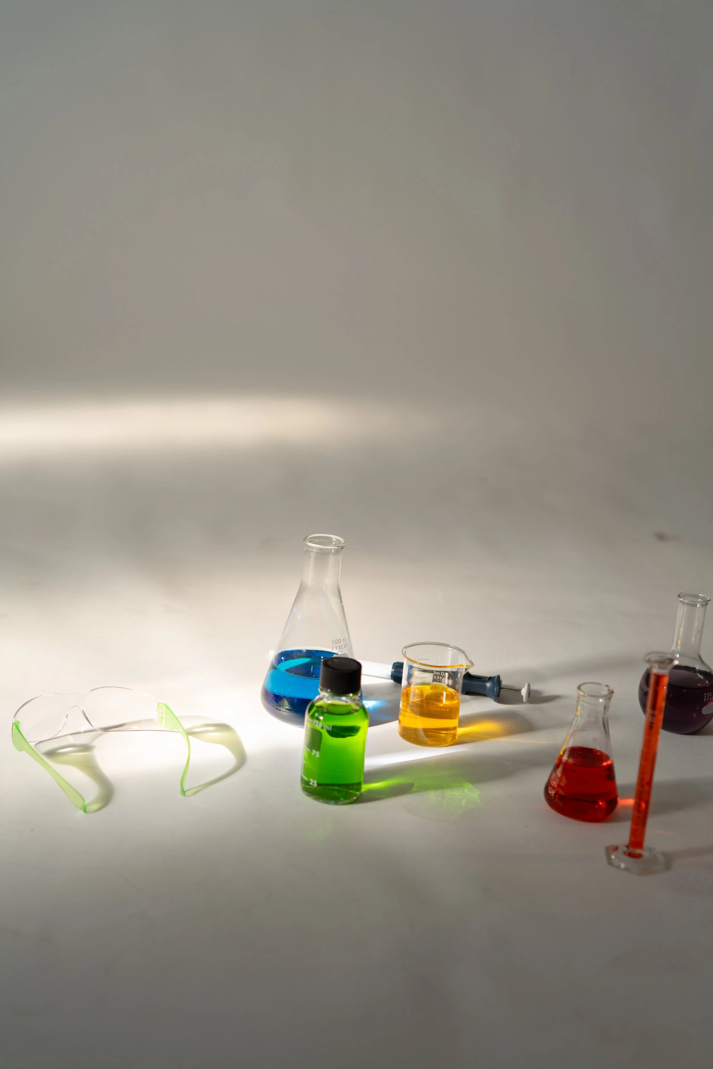 Laboratory glassware with colorful liquids, including a beaker with blue liquid, a conical flask with green liquid, a beaker with yellow liquid, and other containers with red and purple liquids, along with safety goggles.