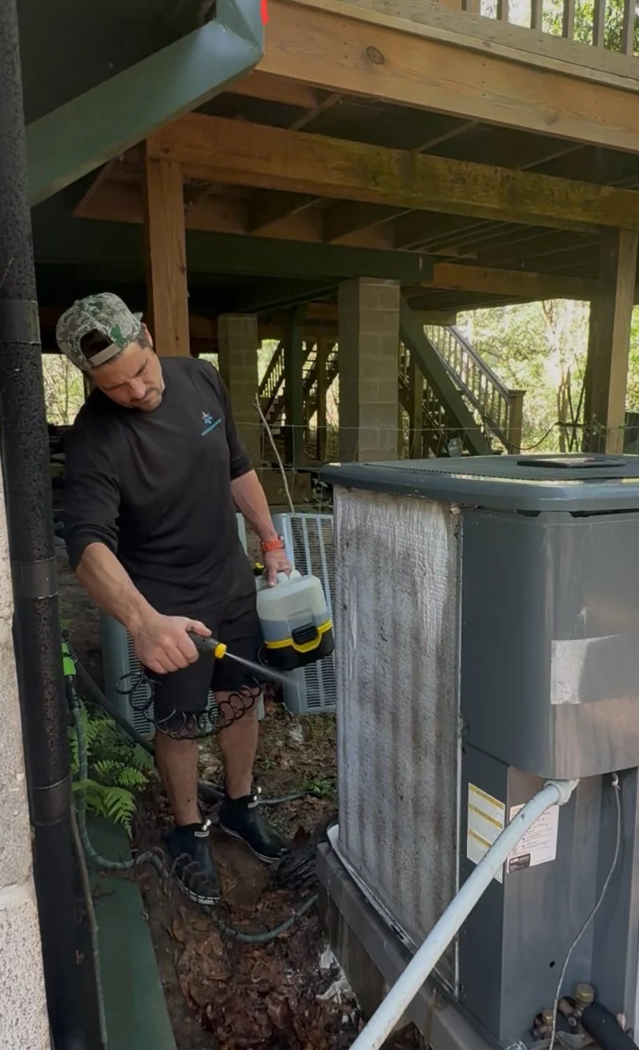 A man in a camouflage cap disinfects an air conditioning unit outside a wooden decked house, wearing a black long sleeve shirt and shorts.