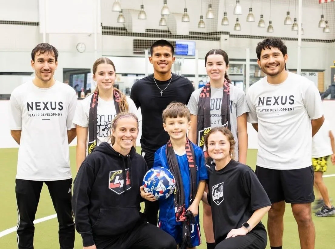 Group of seven people, including kids and adults, smiling on an indoor soccer field. Two children in the front hold a soccer ball, and some are wearing scarves and sports apparel.