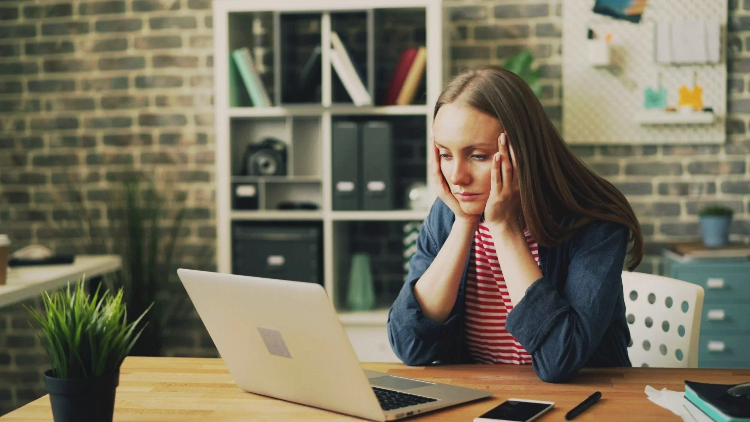 A young woman with brown hair working on a laptop at a desk, looking frustrated or stressed, in a modern office with bookshelves, a potted plant, and organized office supplies.