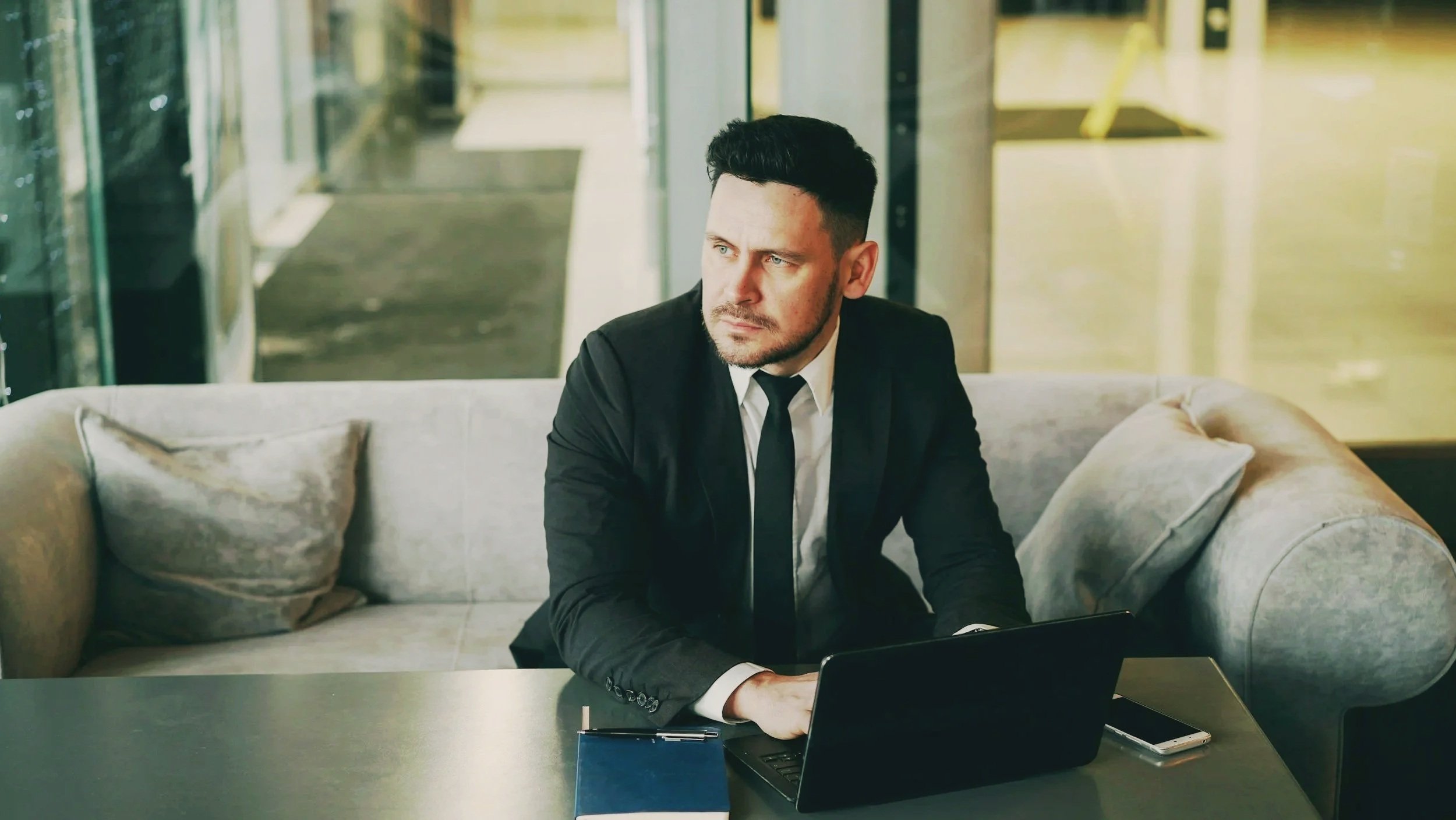 A man in a black suit and tie sitting on a sofa behind a table with a laptop, notebook, and phone, looking contemplative in a modern office lounge.