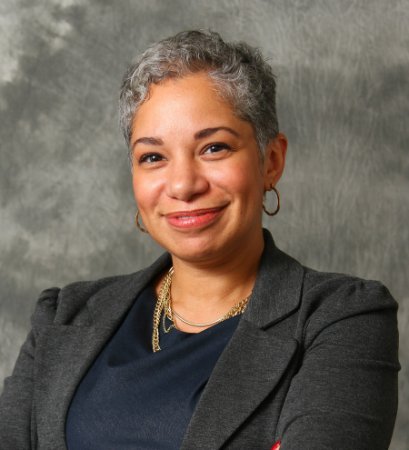 Headshot of Heather Meyer with short gray hair wearing a dark blazer and gold jewelry, posing against a gray background.