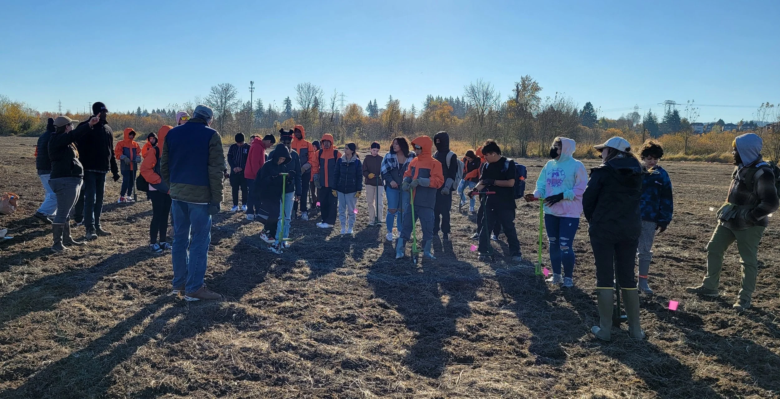 Group of people, including children and adults, gathered outdoors in a field on a sunny day, participating in a planting or environmental activity.