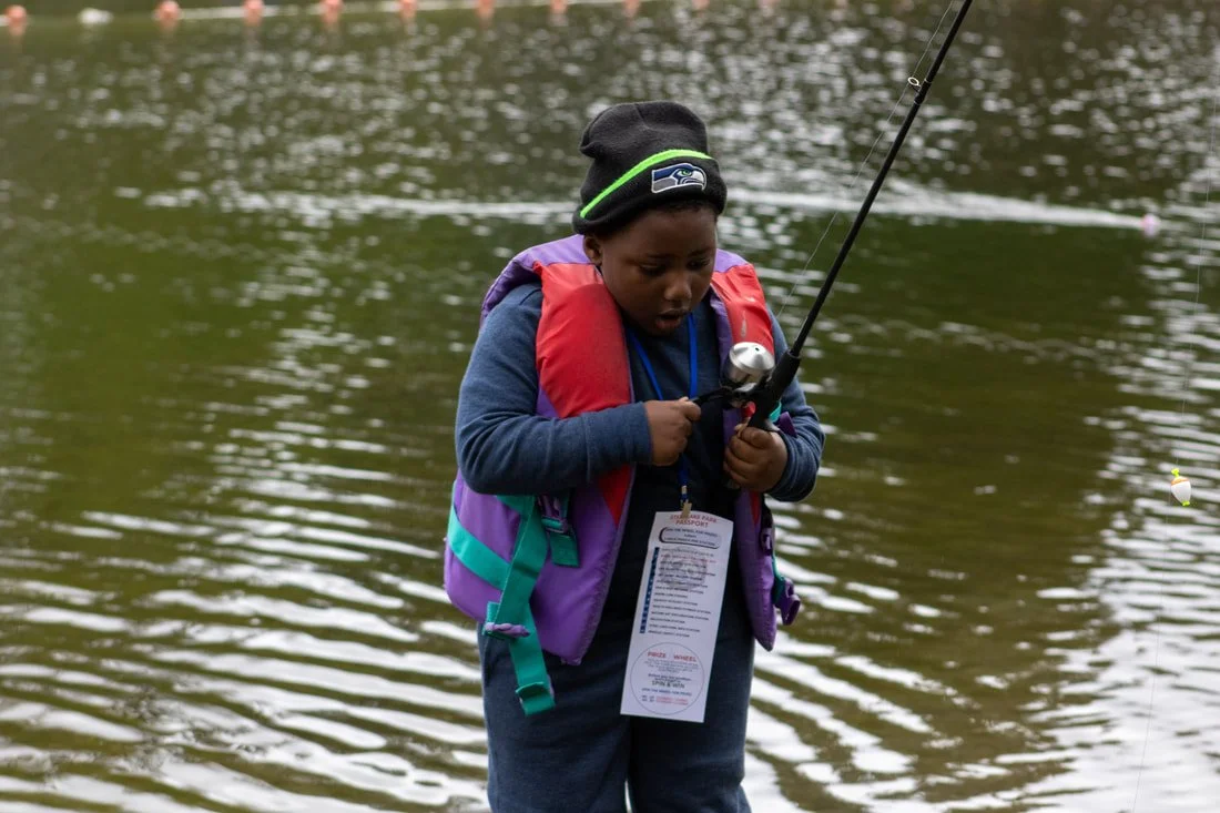 A young boy wearing a black beanie, blue jacket, and purple and red life jacket, standing by a river and holding a fishing rod, concentrating on fishing.