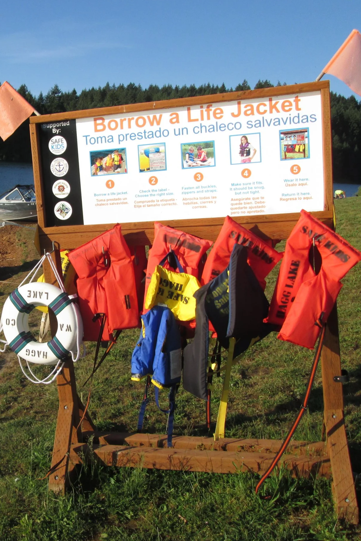 A sign with instructions on how to borrow a life jacket at a lakeside, with multiple life jackets hanging on a wooden stand, and a boat visible in the background.